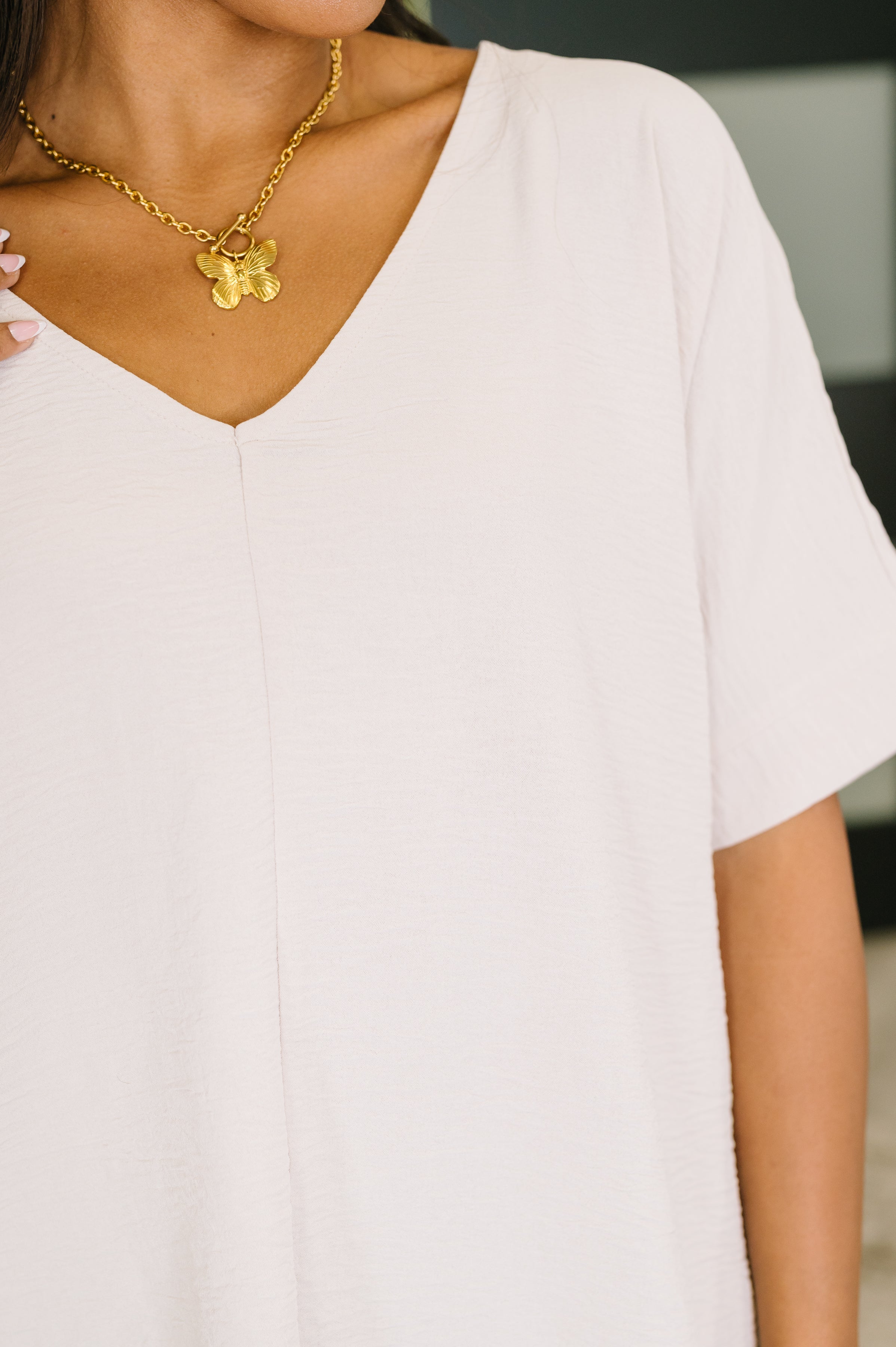 Close-up of woman wearing sand beige crinkle textured V-neck top with short sleeves and gold butterfly pendant necklace