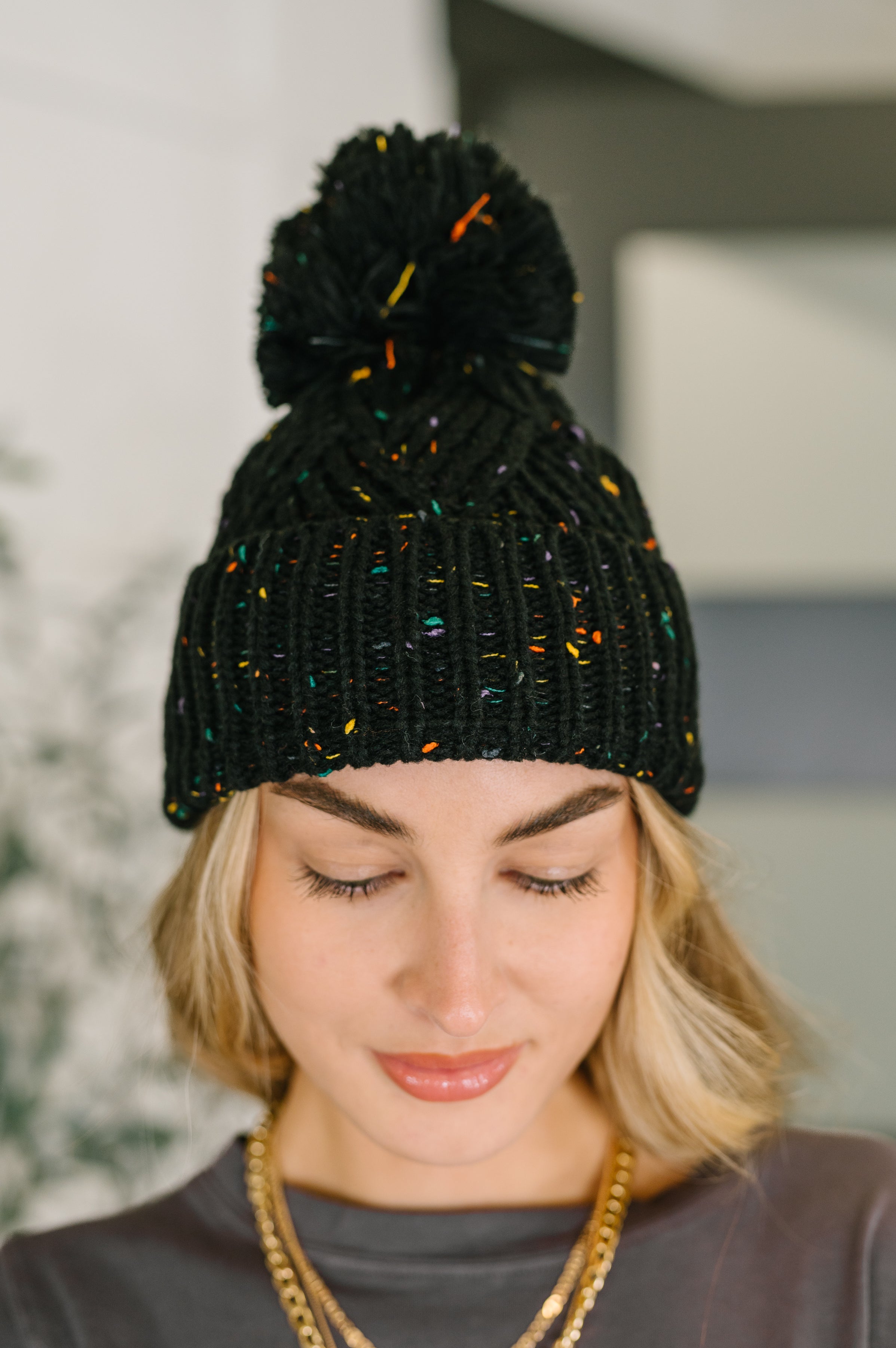 Close-up of woman wearing black knit beanie with colorful specks and large pom pom on top indoors