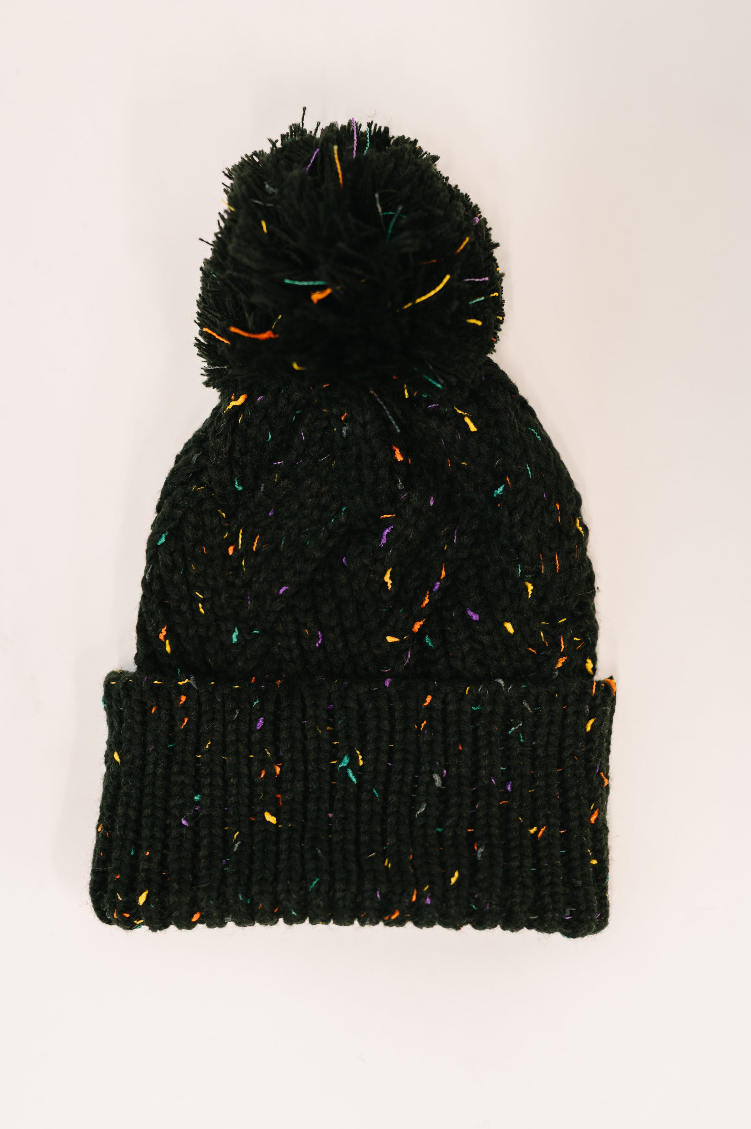 Black knit beanie with multicolored flecks and large pom pom on top laid flat on white background