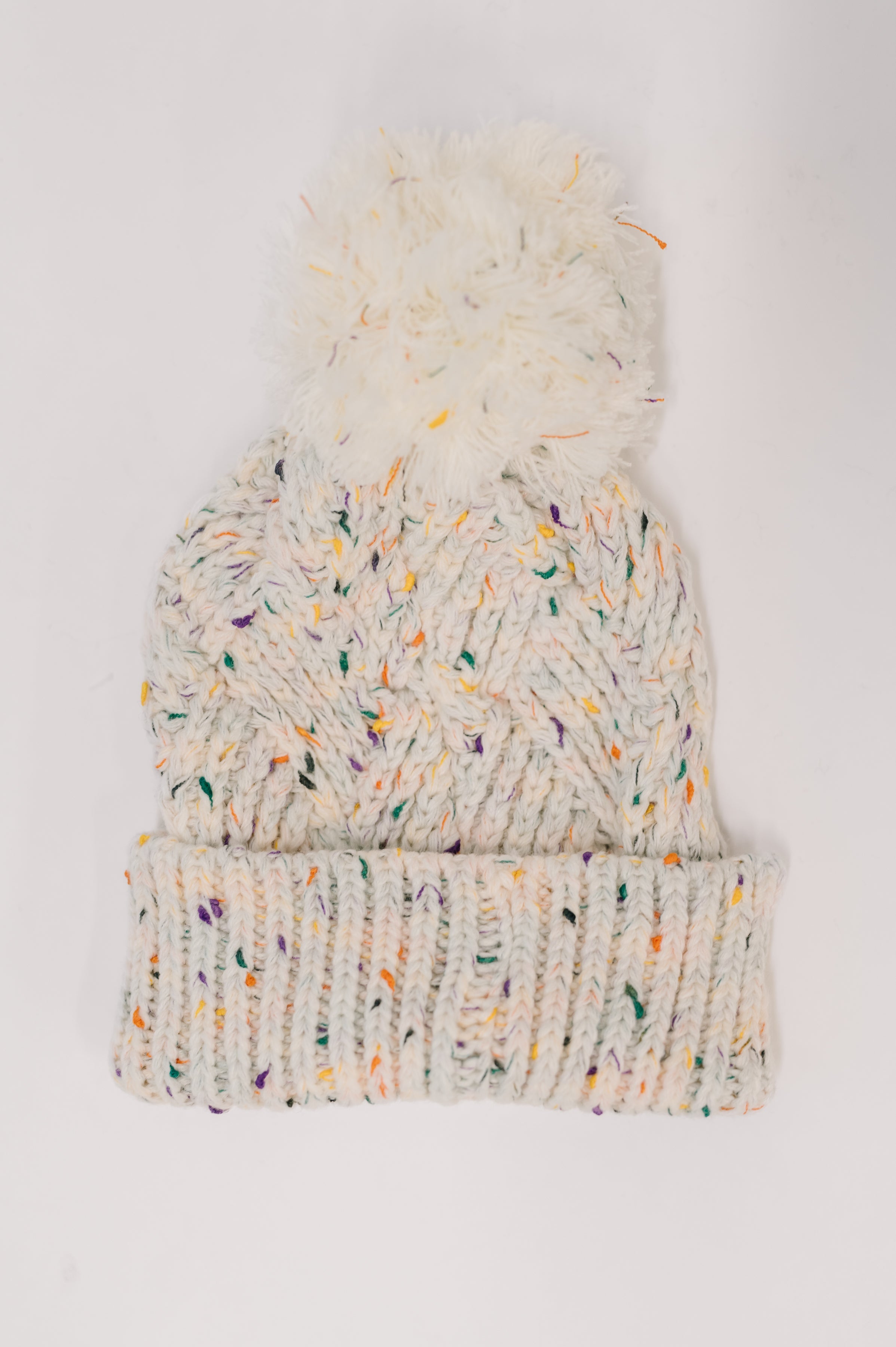 Ivory knit beanie with multicolored flecks and large fluffy pom pom on top laid flat on light background