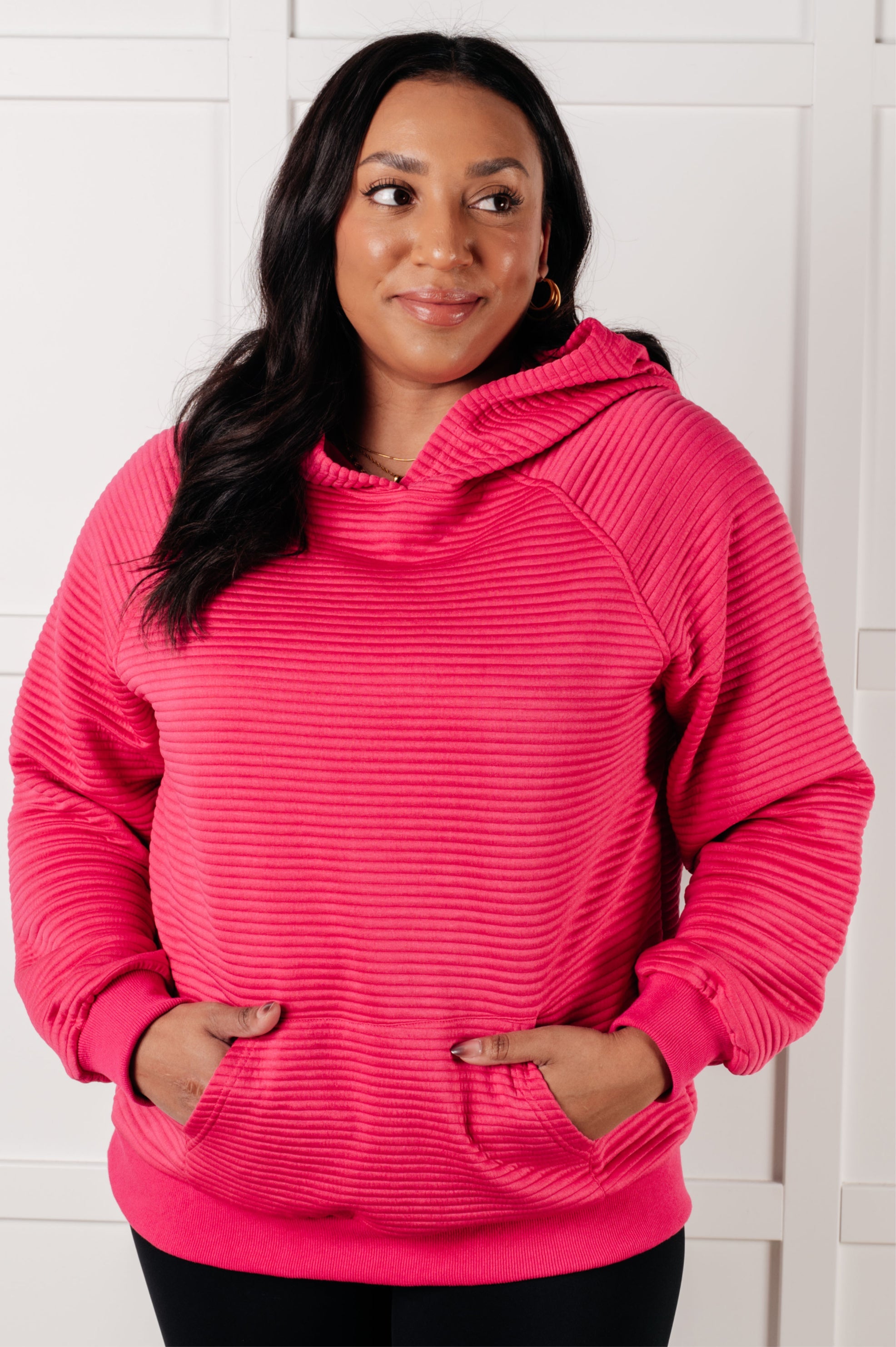 Plus size woman with dark hair wearing bright rose textured crosswalk hoodie with hands in front pocket indoors