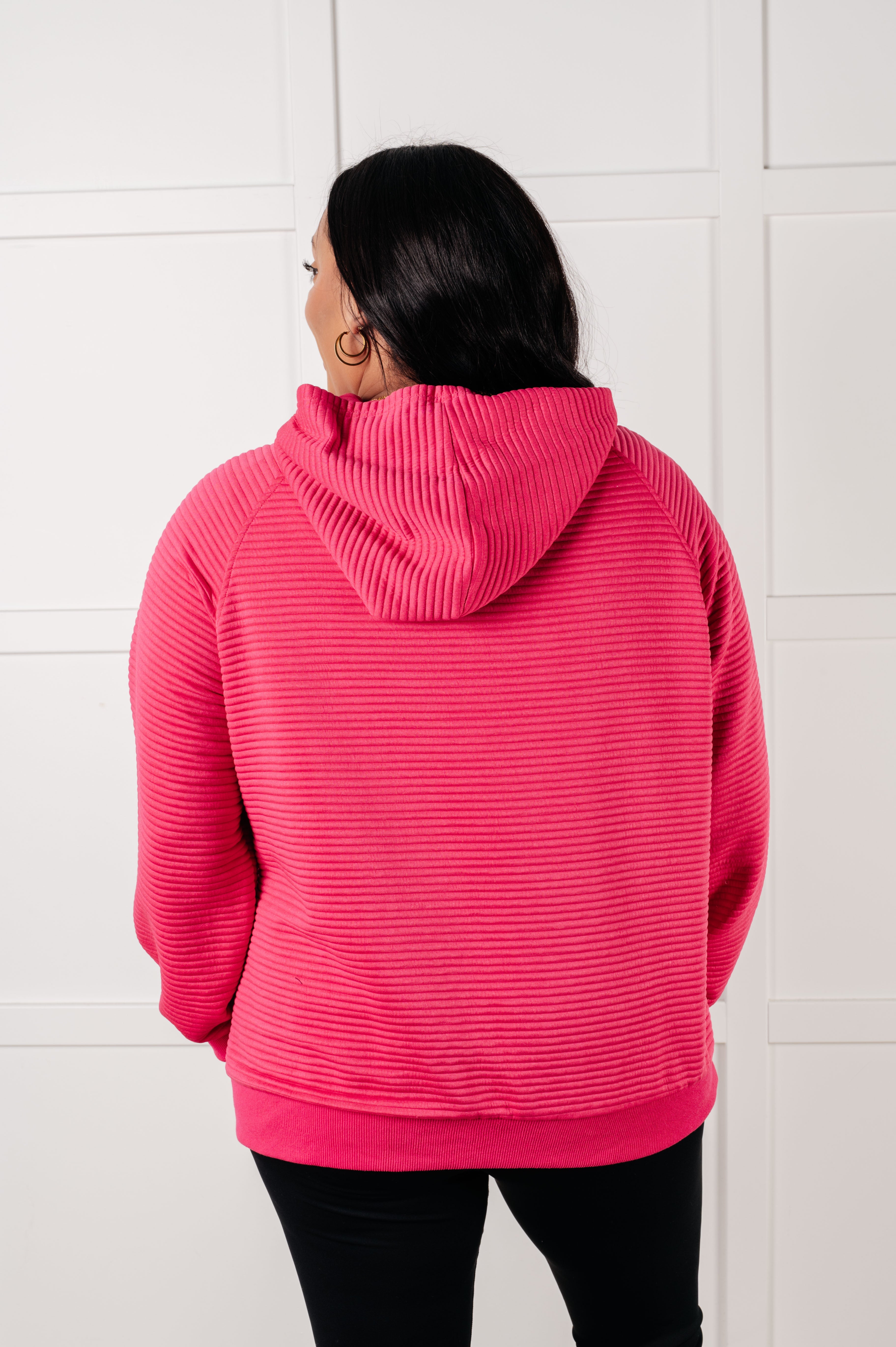 Back view of woman wearing rose pink ribbed textured hoodie with hood down standing indoors