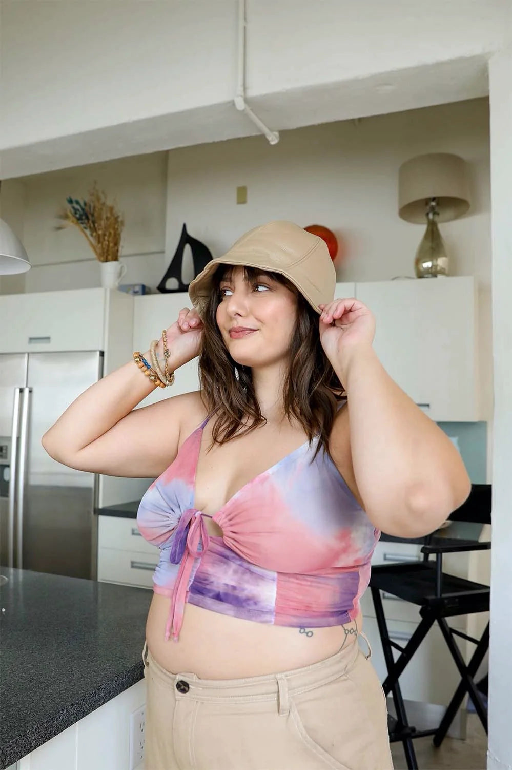 Plus size woman adjusting beige bucket hat wearing pastel tie dye brami top with front tie detail and beige pants in kitchen