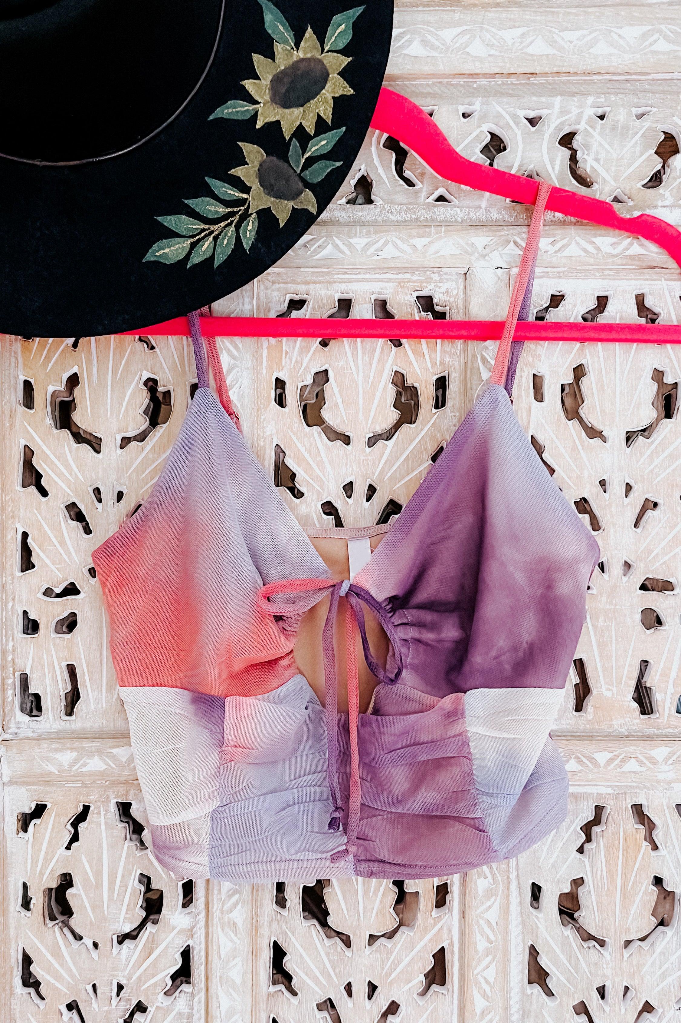 Purple, pink, and orange tie dye mesh brami top with front tie detail hanging on carved wooden screen with black hat