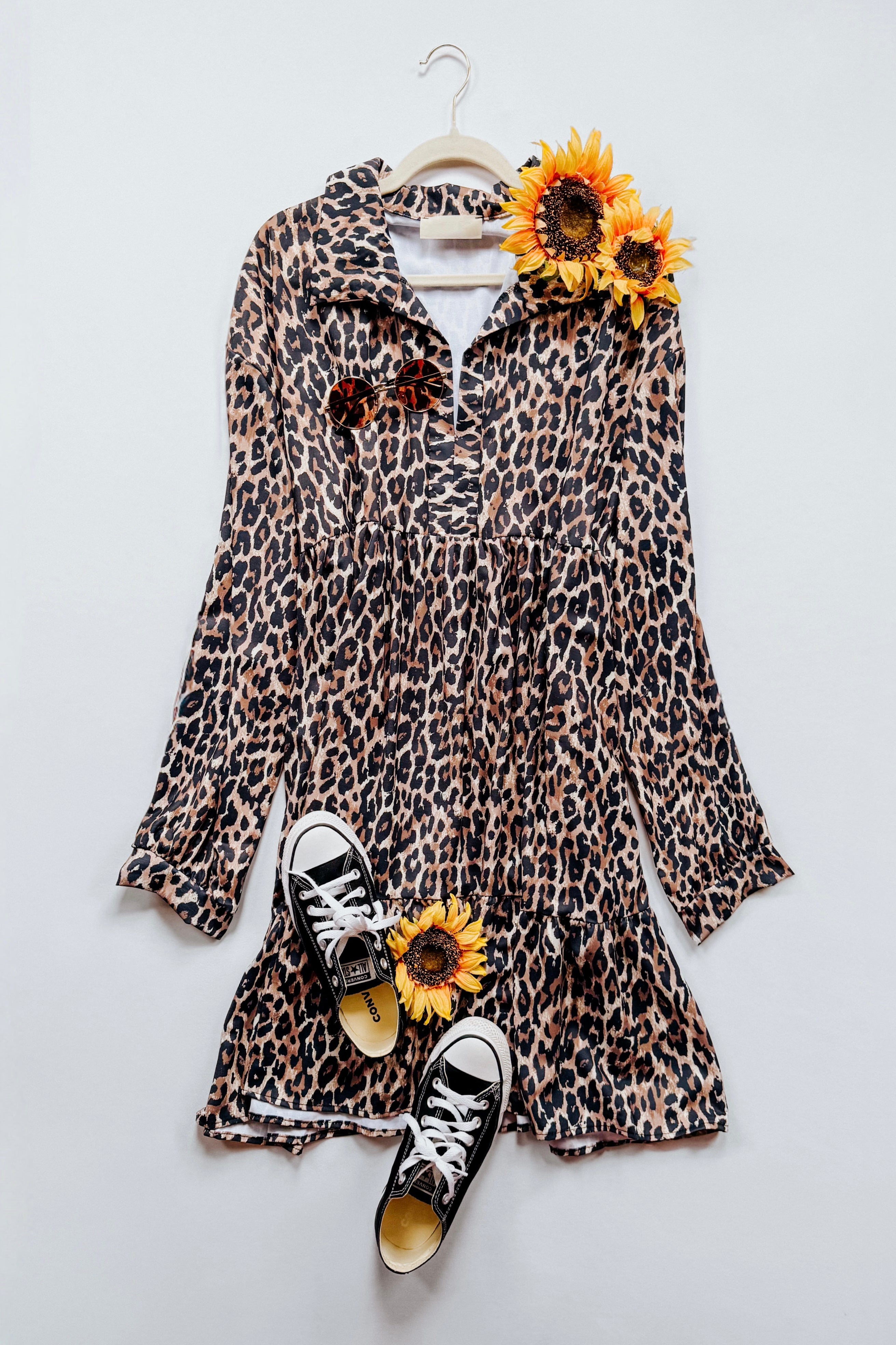 Long-sleeve babydoll dress in leopard print displayed flat with black sneakers and sunflowers on white background