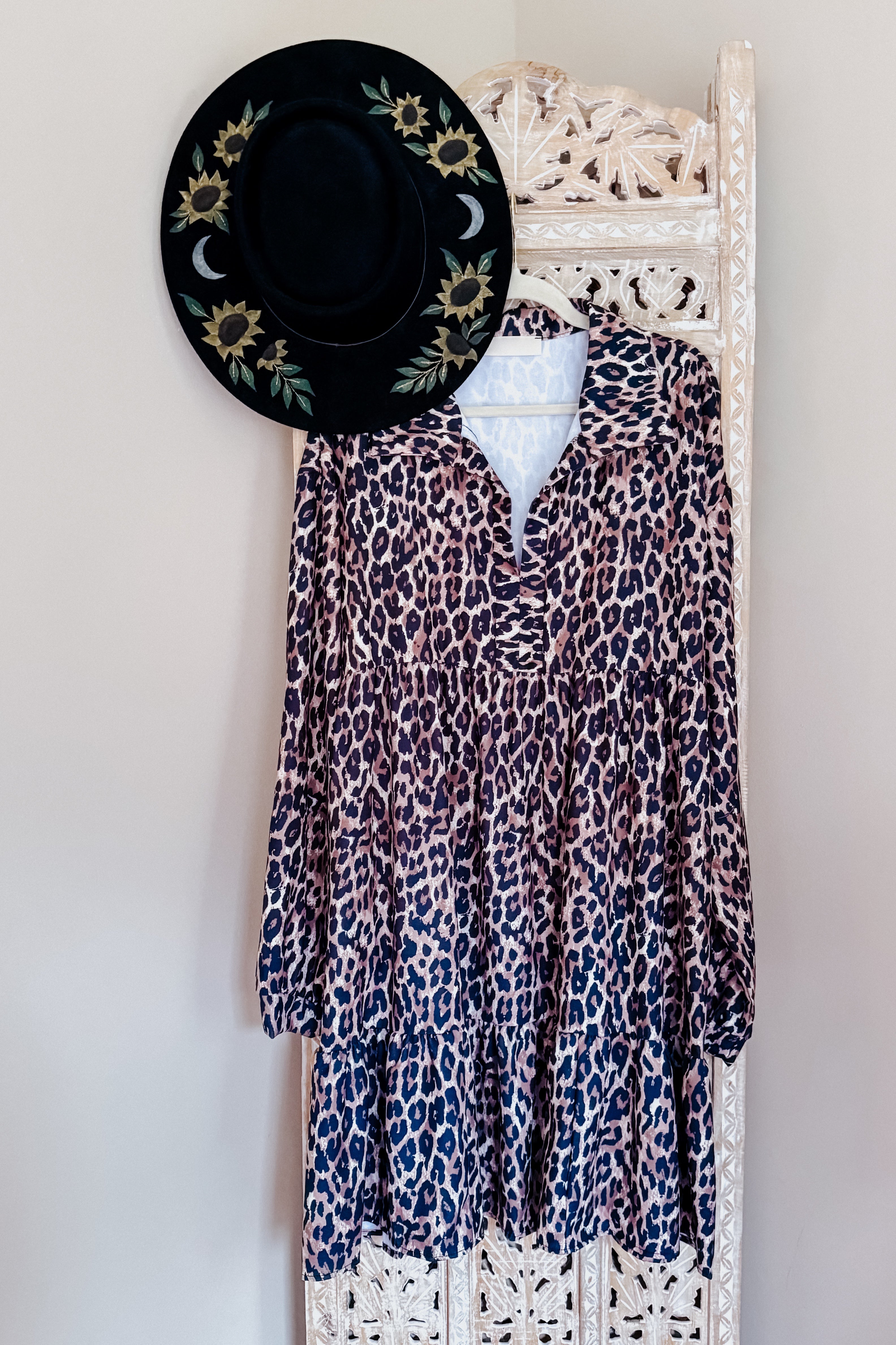 Leopard print long-sleeve babydoll dress hanging on carved wooden screen with black sunflower hat above it