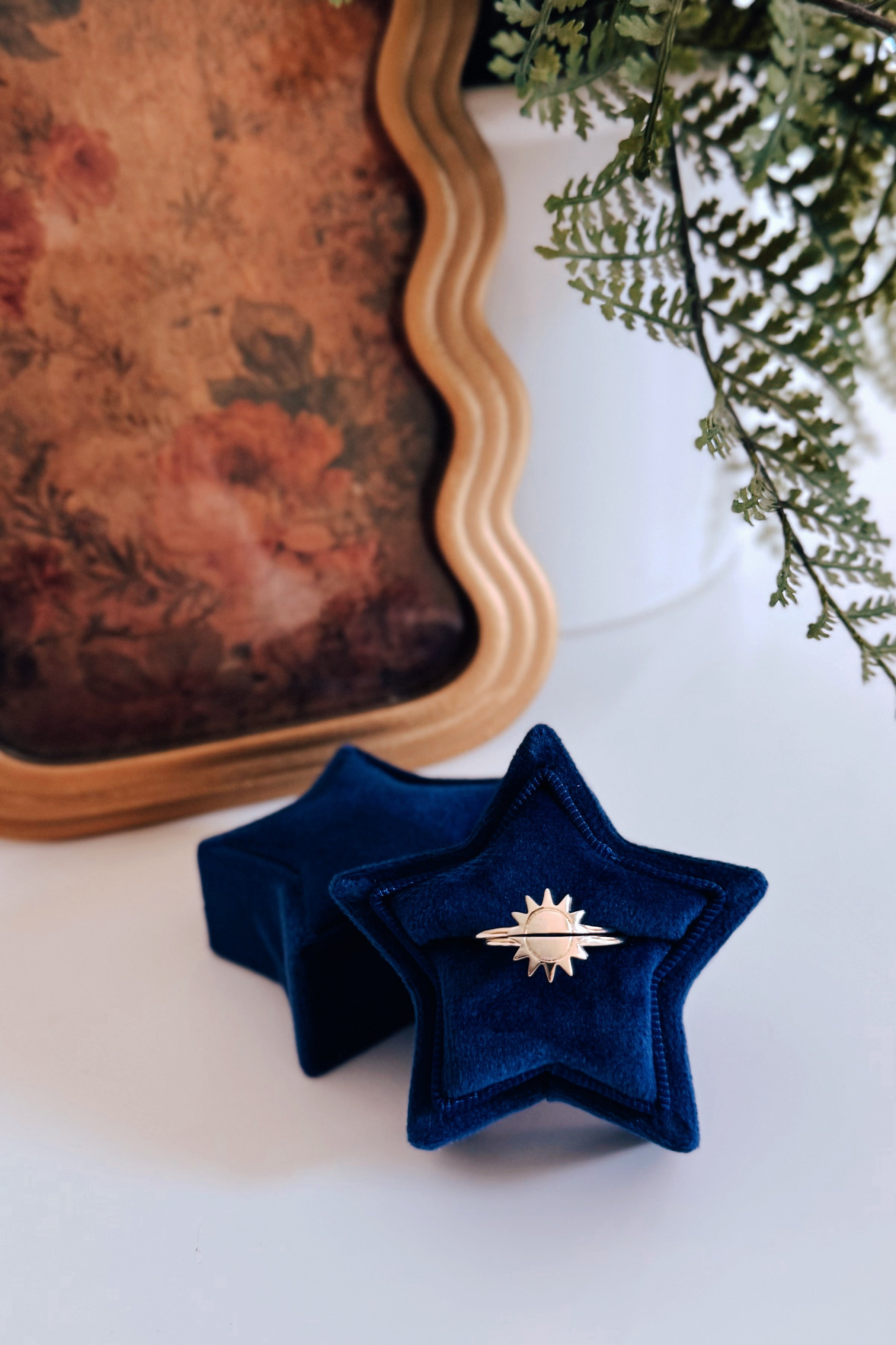 Gold sun-shaped ring duo displayed on star-shaped navy velvet ring box with vintage floral artwork and greenery nearby