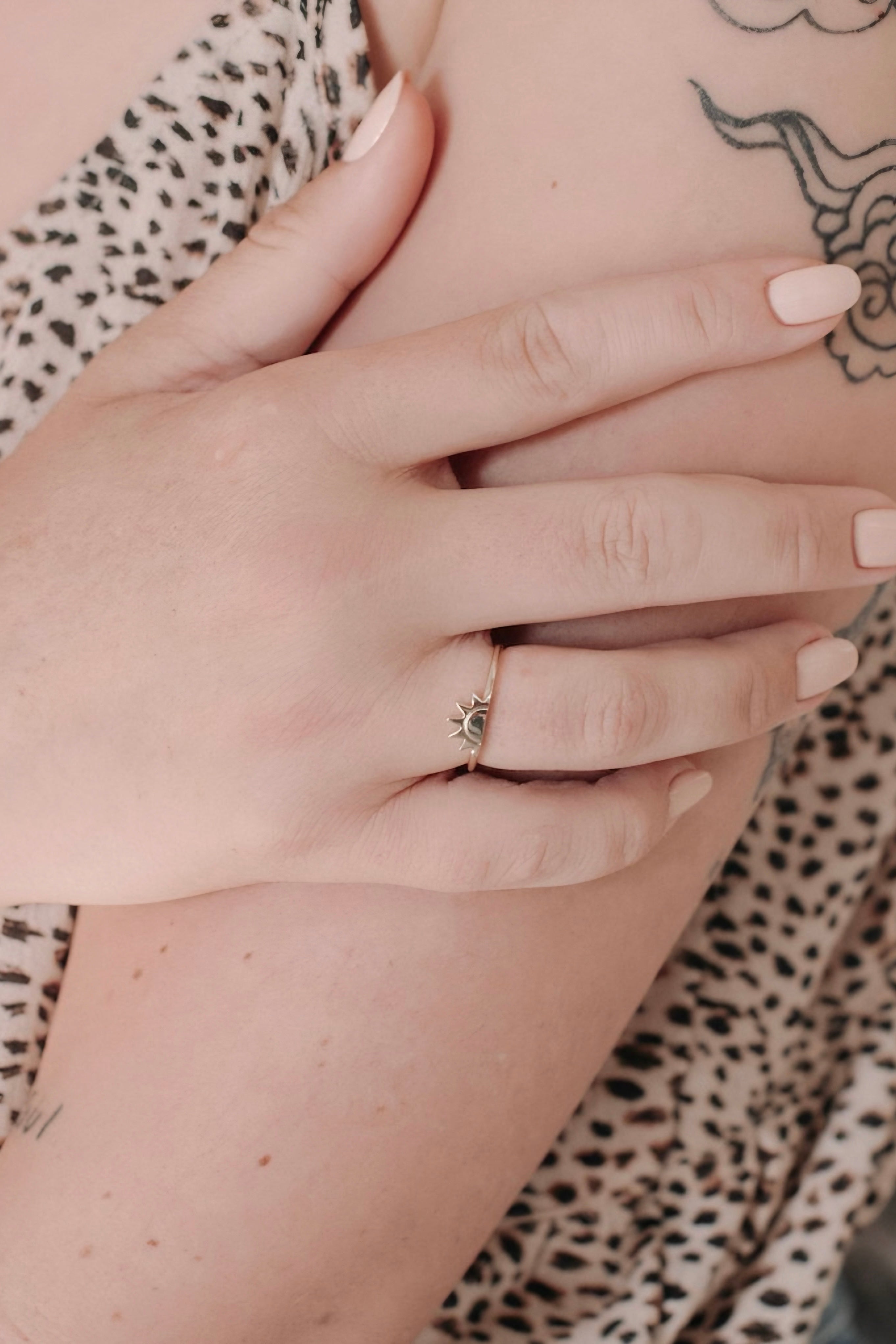 Hand wearing delicate gold sun ring resting on crossed arms with leopard print fabric and tattoo details visible