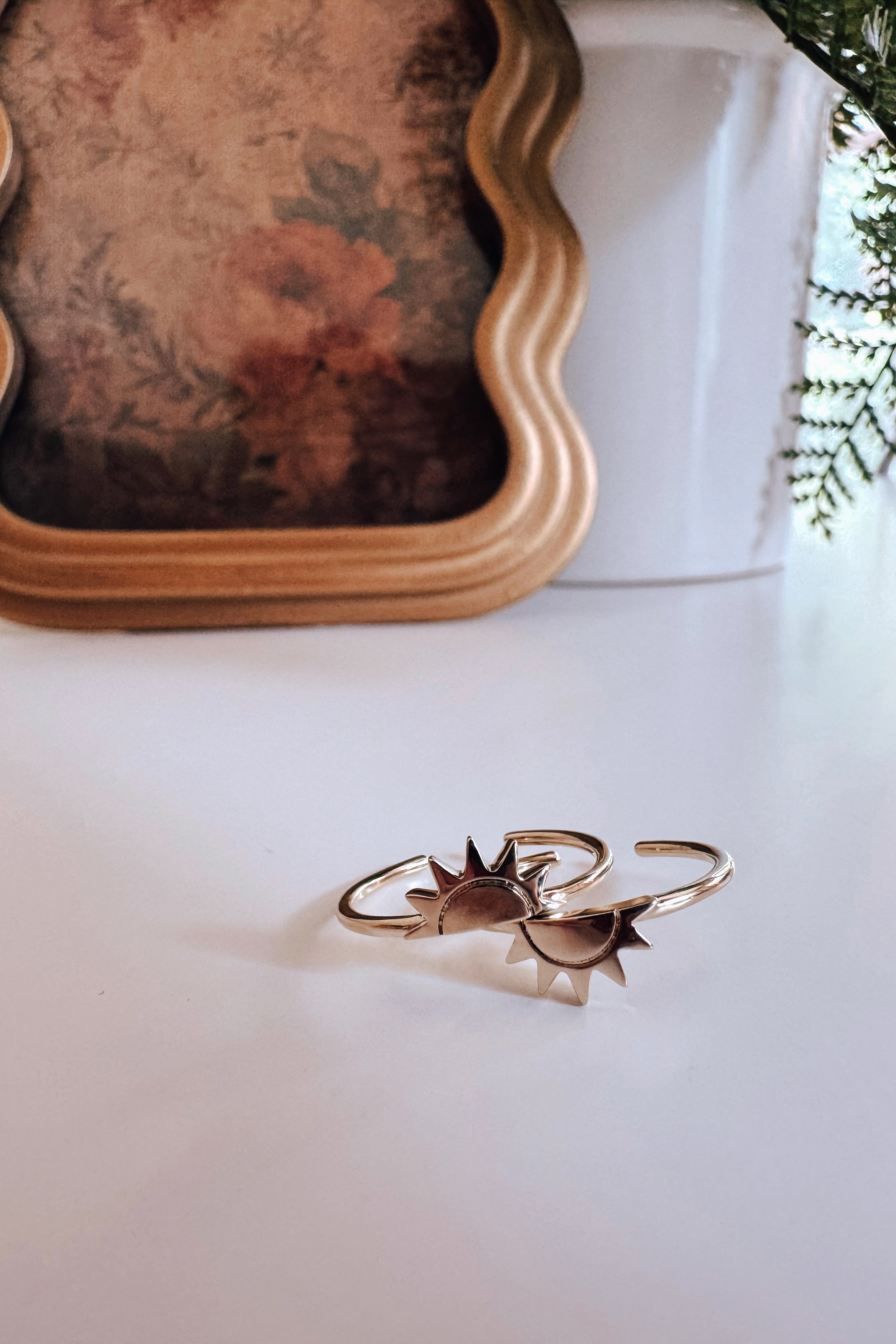 Two gold open-ended rings with sunburst designs resting on white surface near gold frame and greenery