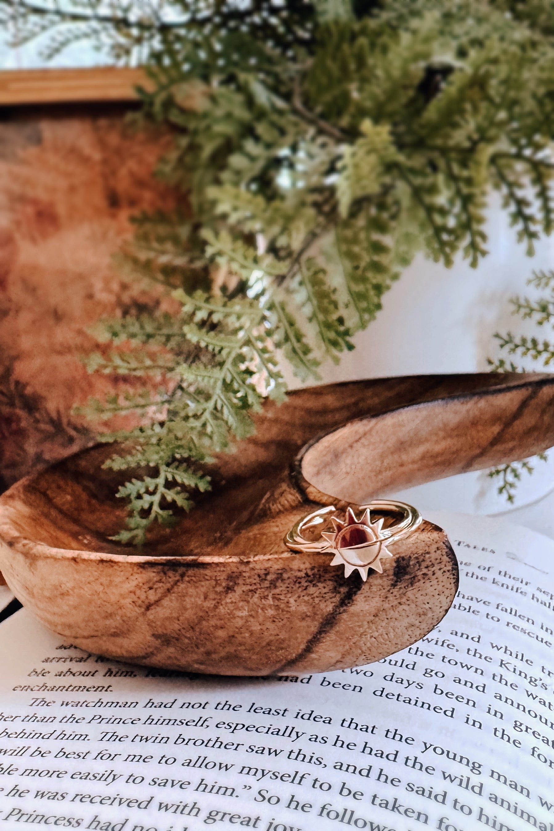 Gold sun-shaped ring resting in carved wooden bowl beside open book pages and green fern leaves