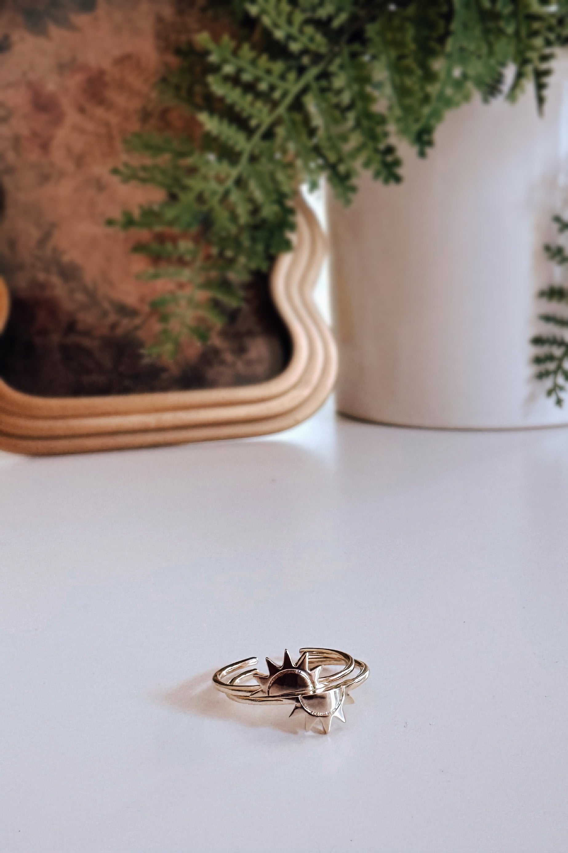 Gold double ring featuring sun and crescent moon designs placed on white surface with plants and mirror in background