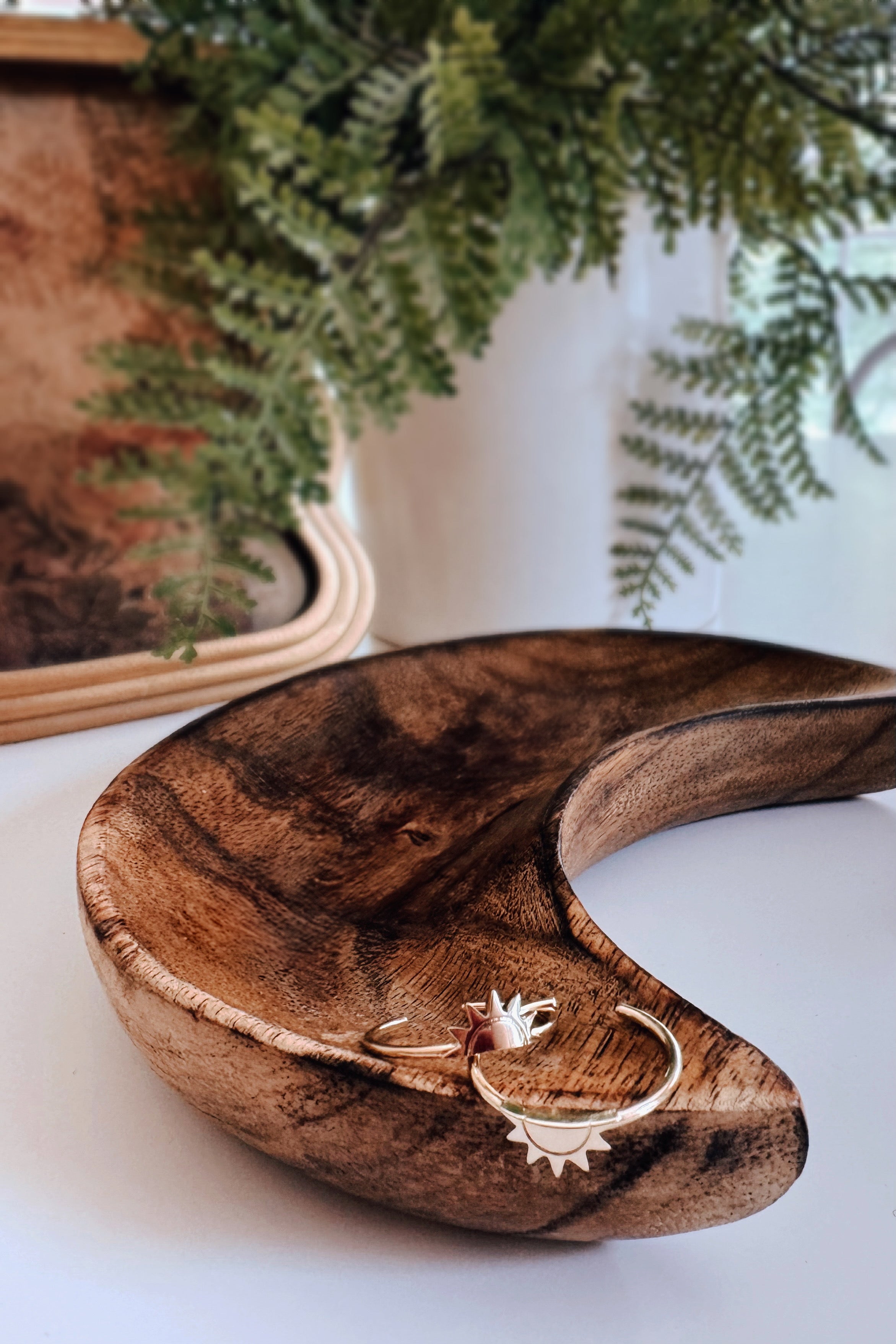 Two delicate gold rings with sun designs displayed on a crescent-shaped wooden dish with greenery backdrop