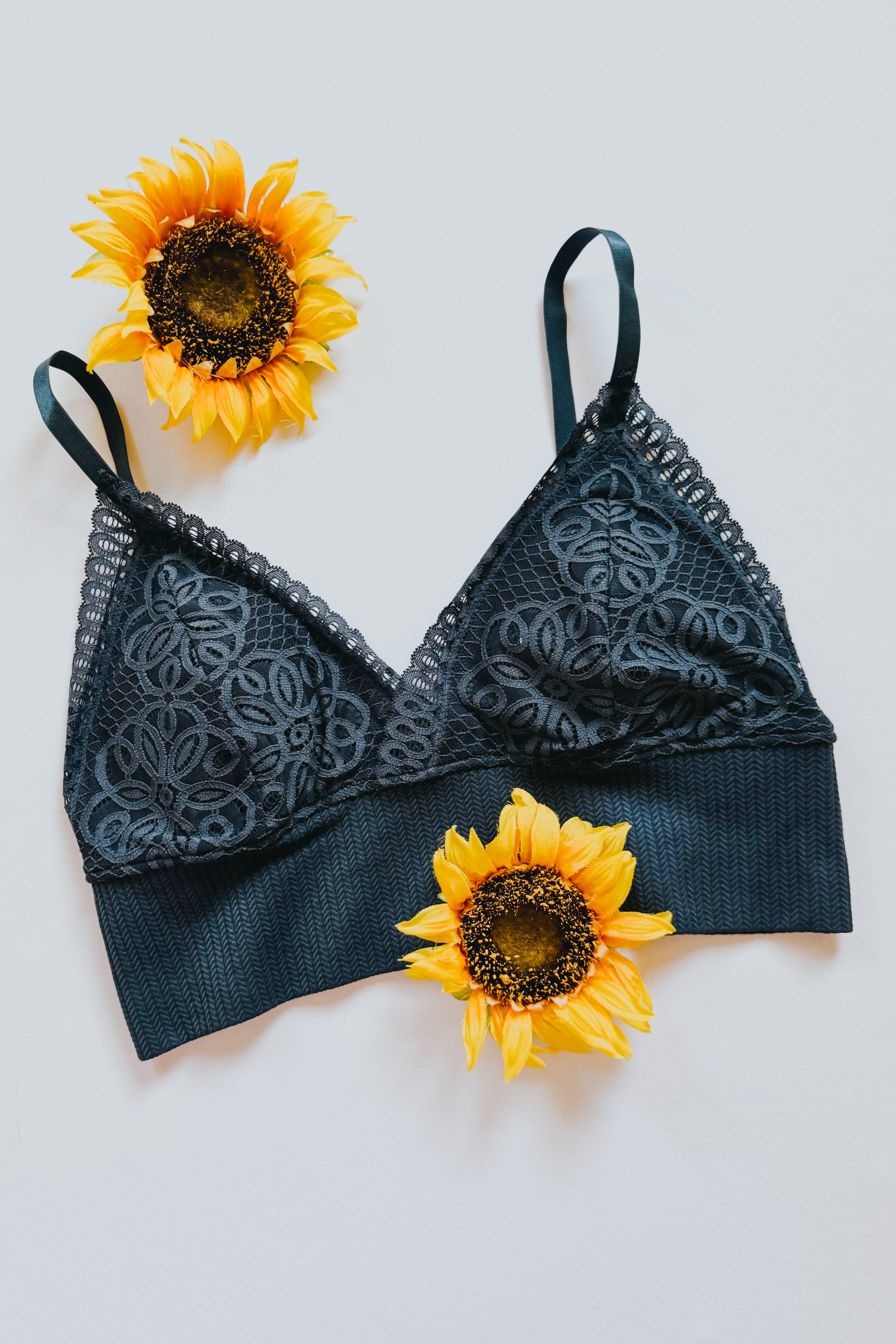 Black lace bralette with floral mesh design and textured band styled flat with two bright sunflowers on light background