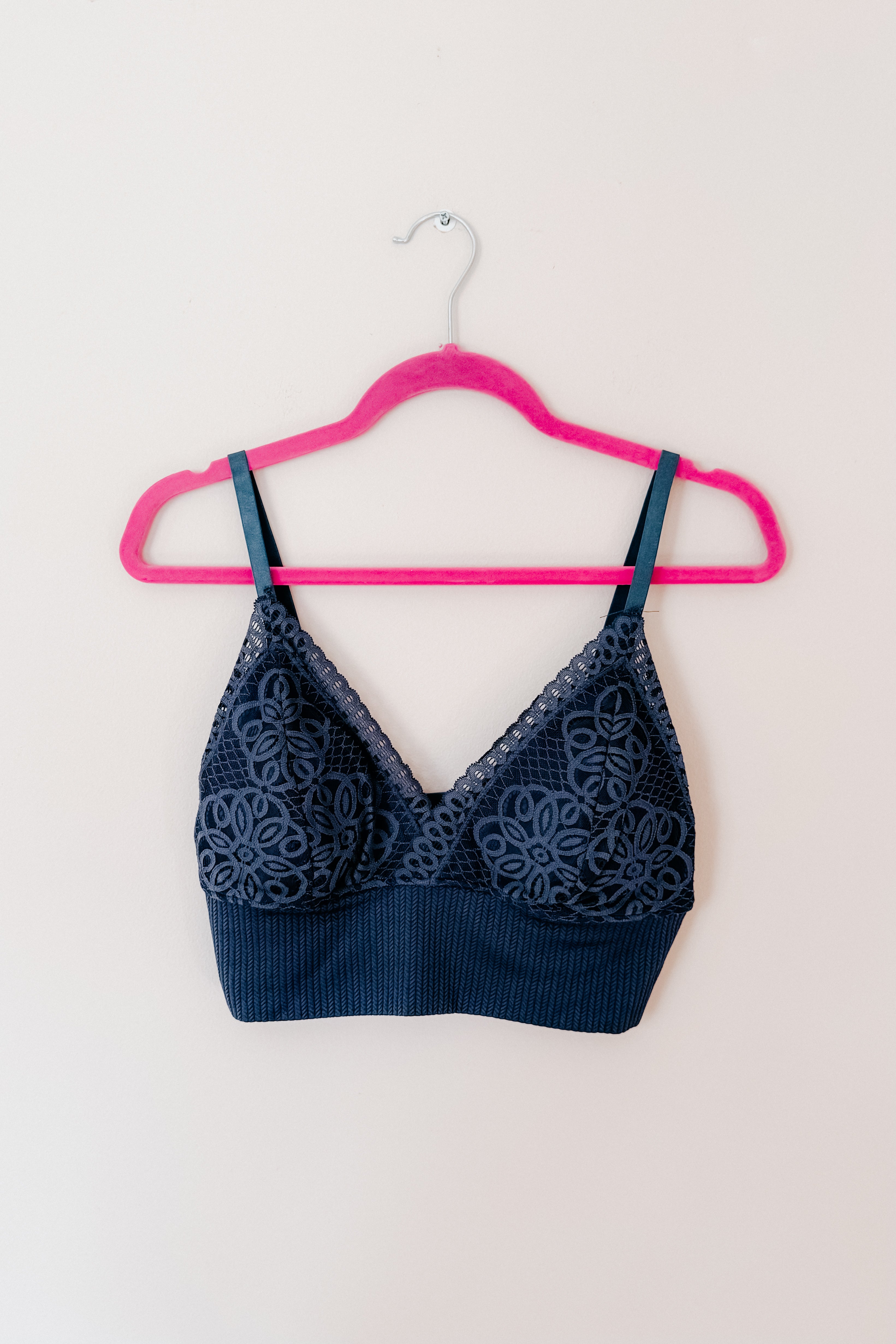 Navy lace bralette with floral patterns and ribbed underband hanging on bright pink hanger against white wall