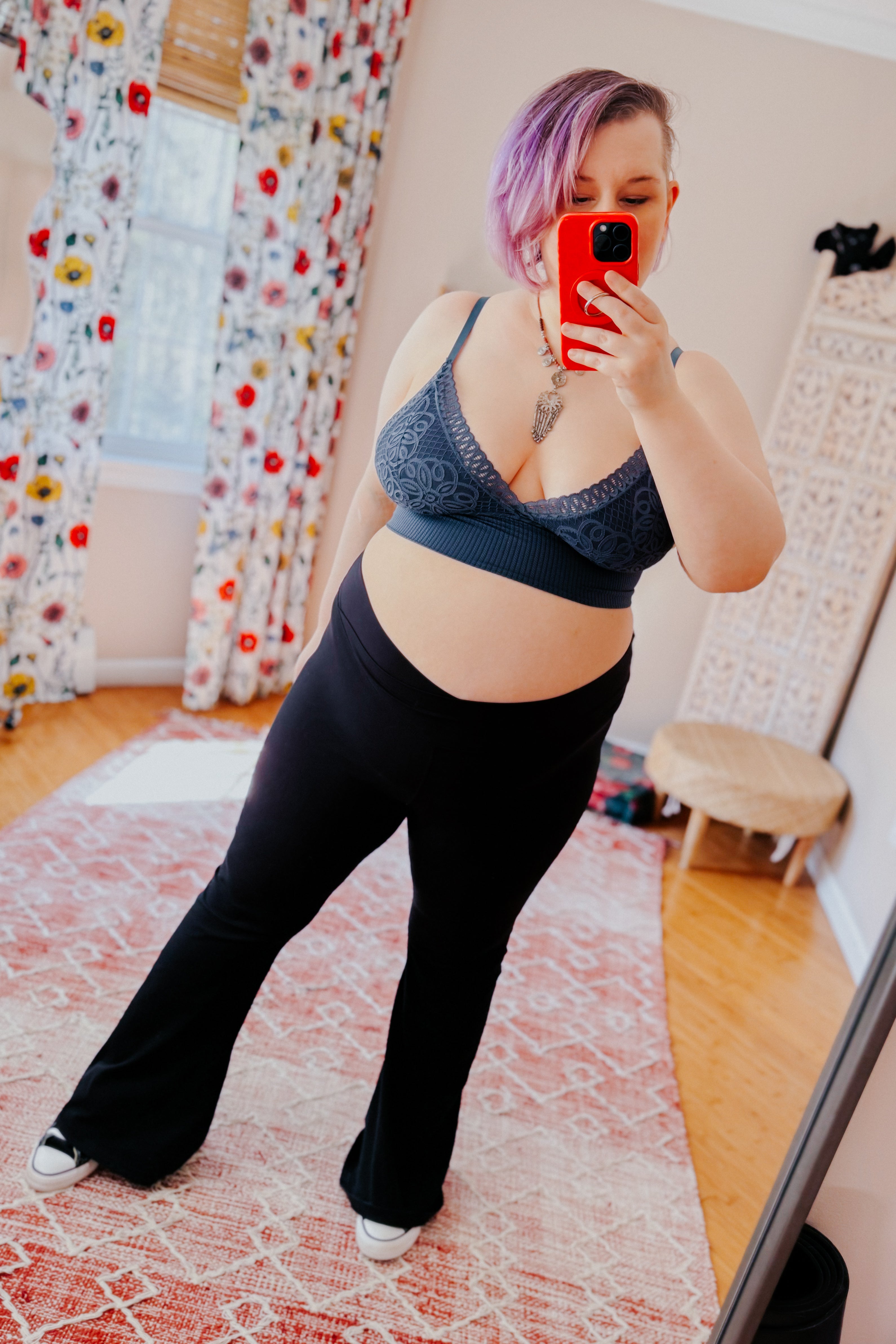Woman with purple hair wearing dusty blue lace Daydream bralette and black pants taking mirror selfie in bright room