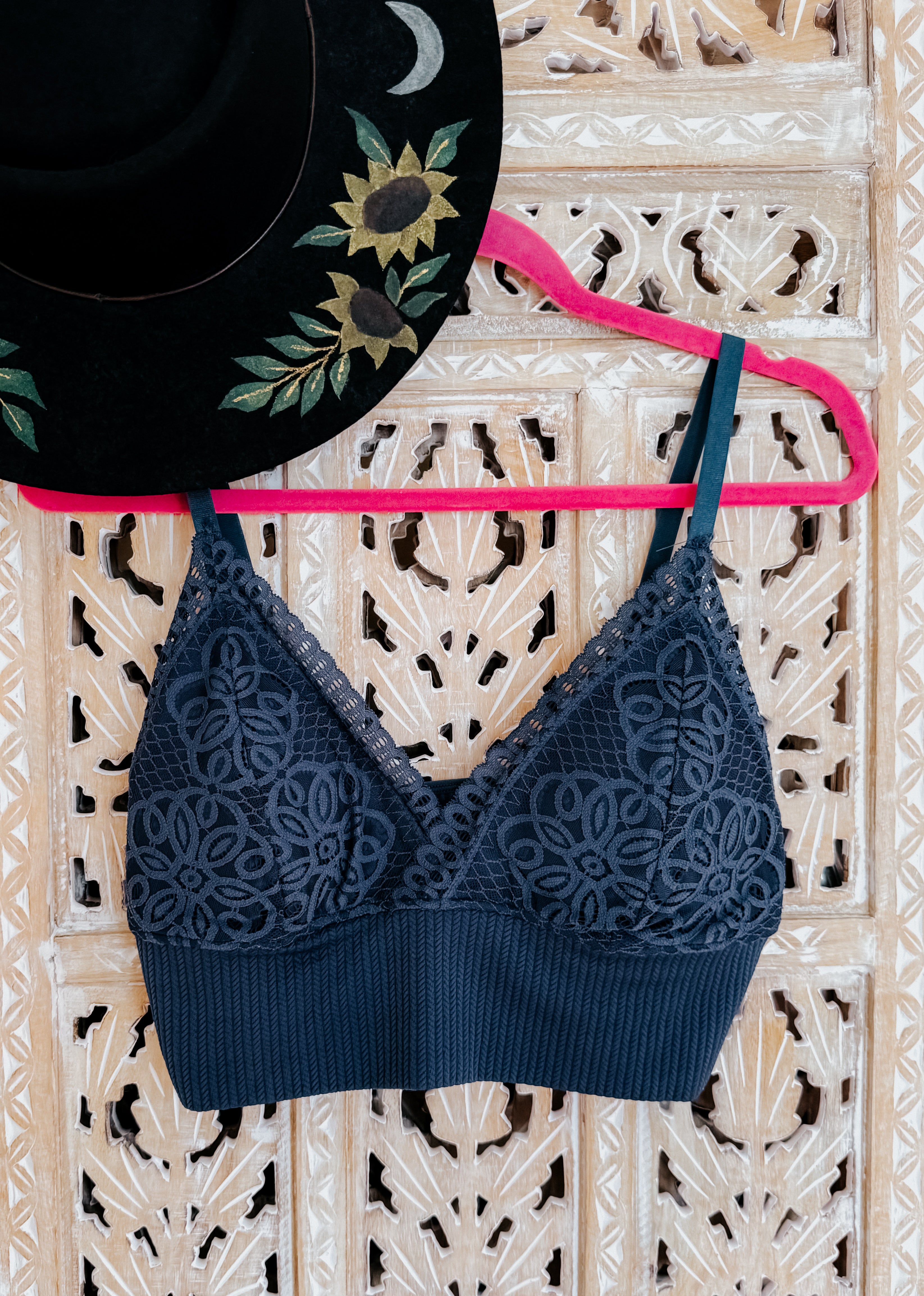 Black lace bralette with floral mesh cups and ribbed band hanging on pink hanger against wooden carved screen