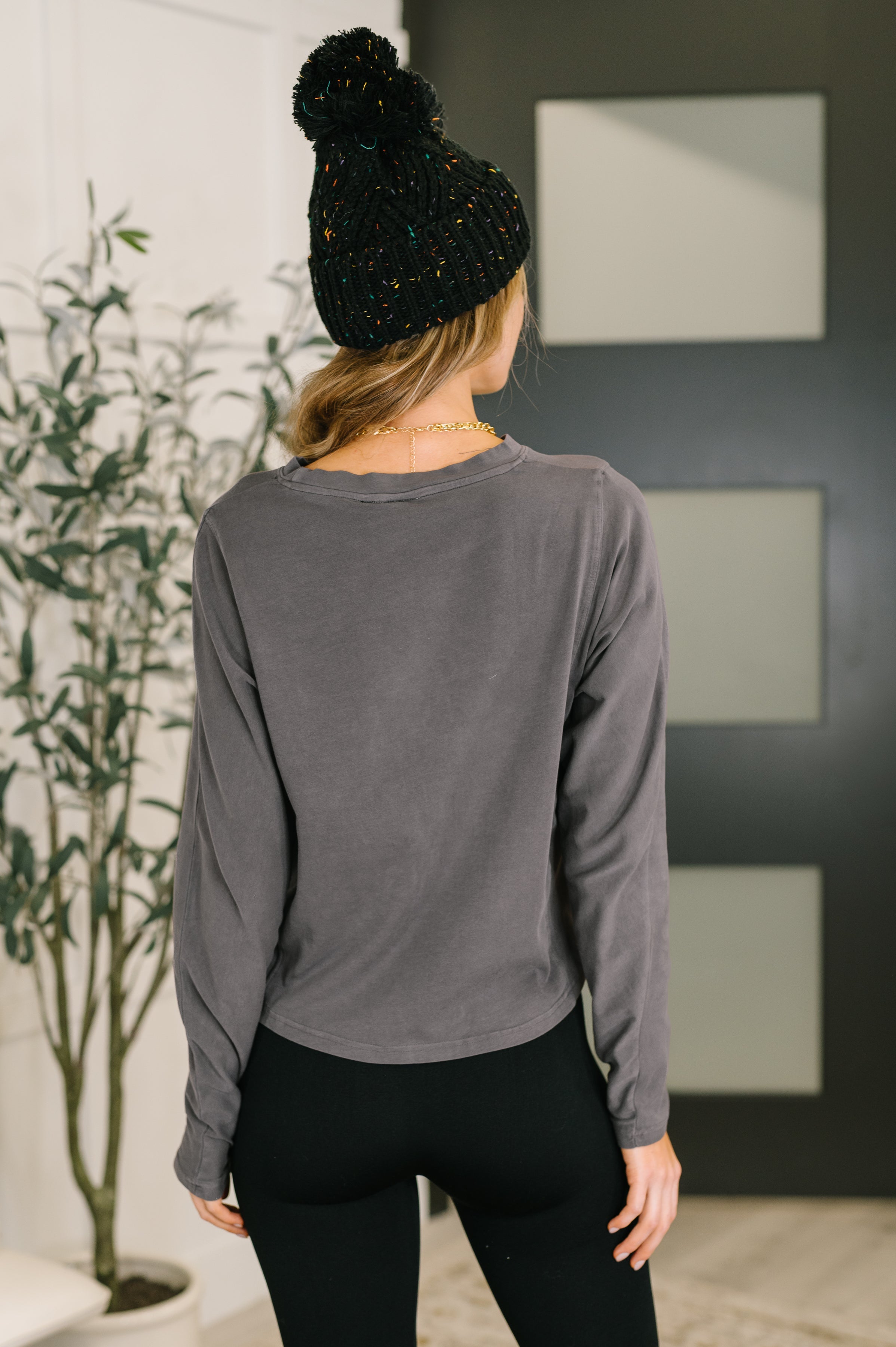 Woman wearing gray dedicated dolman sleeve top and black leggings standing indoors with knitted hat on blonde hair