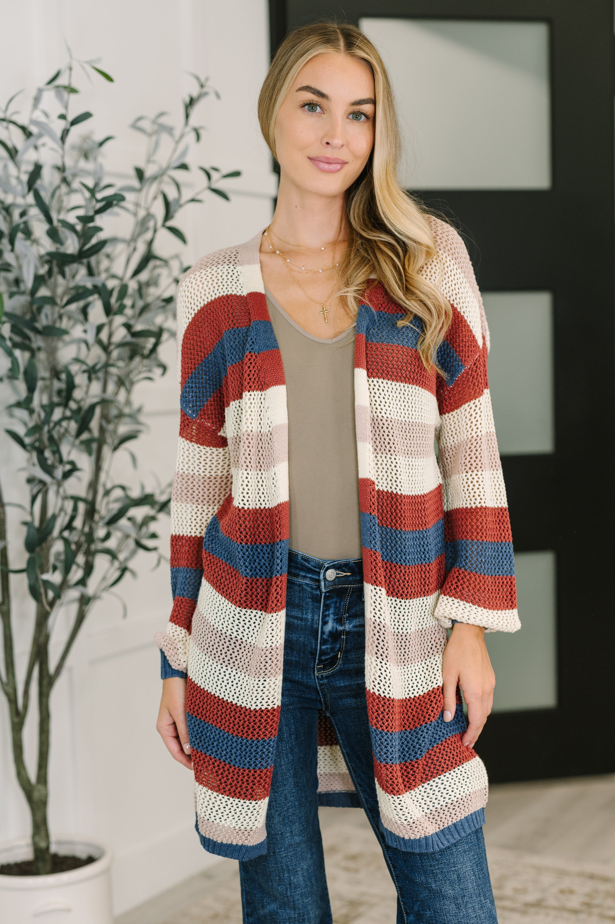 Woman wearing open striped knit cardigan in red, blue, cream, and beige over taupe top and blue jeans indoors