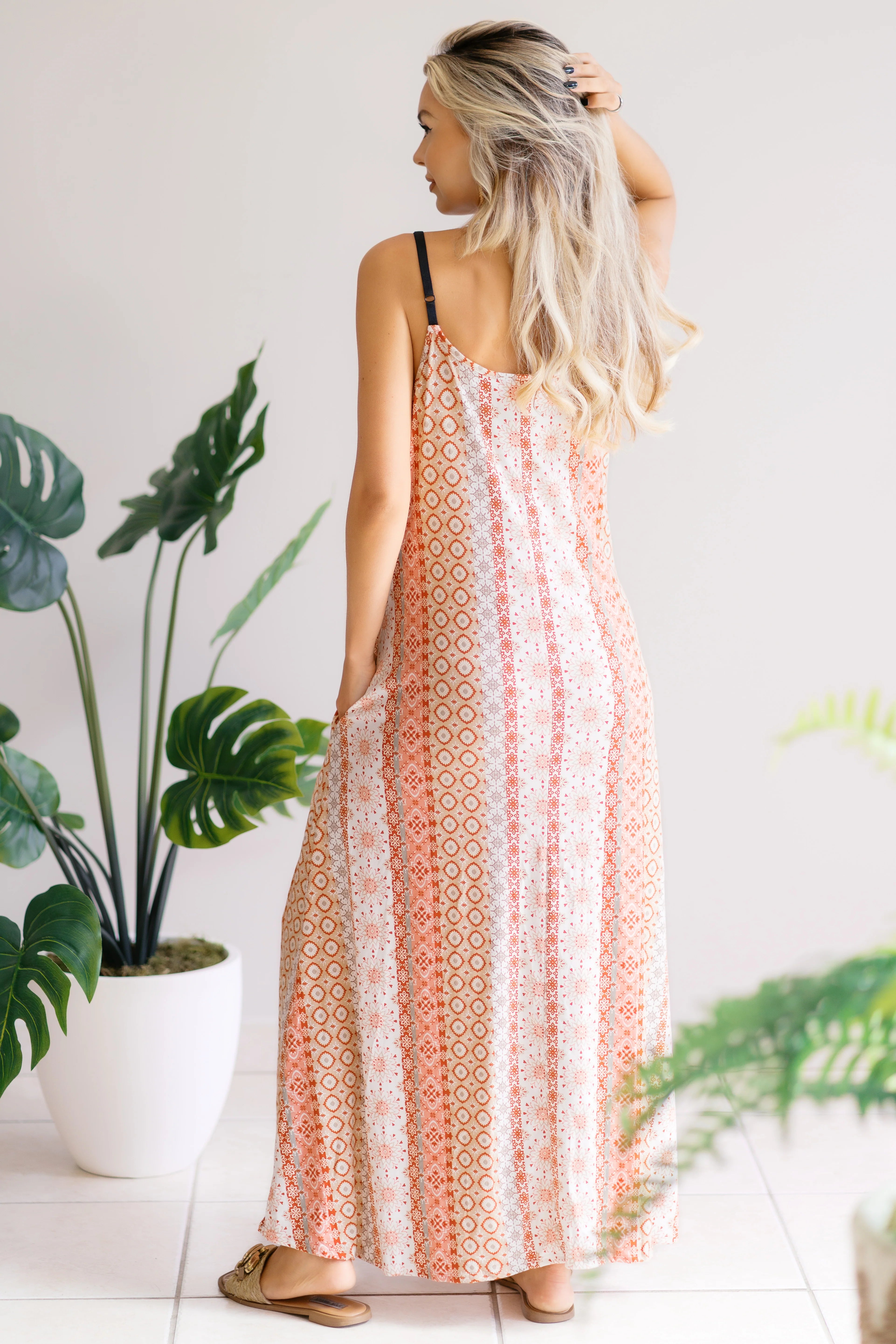 Woman with long blonde hair wearing sleeveless patterned maxi dress standing indoors near potted plants
