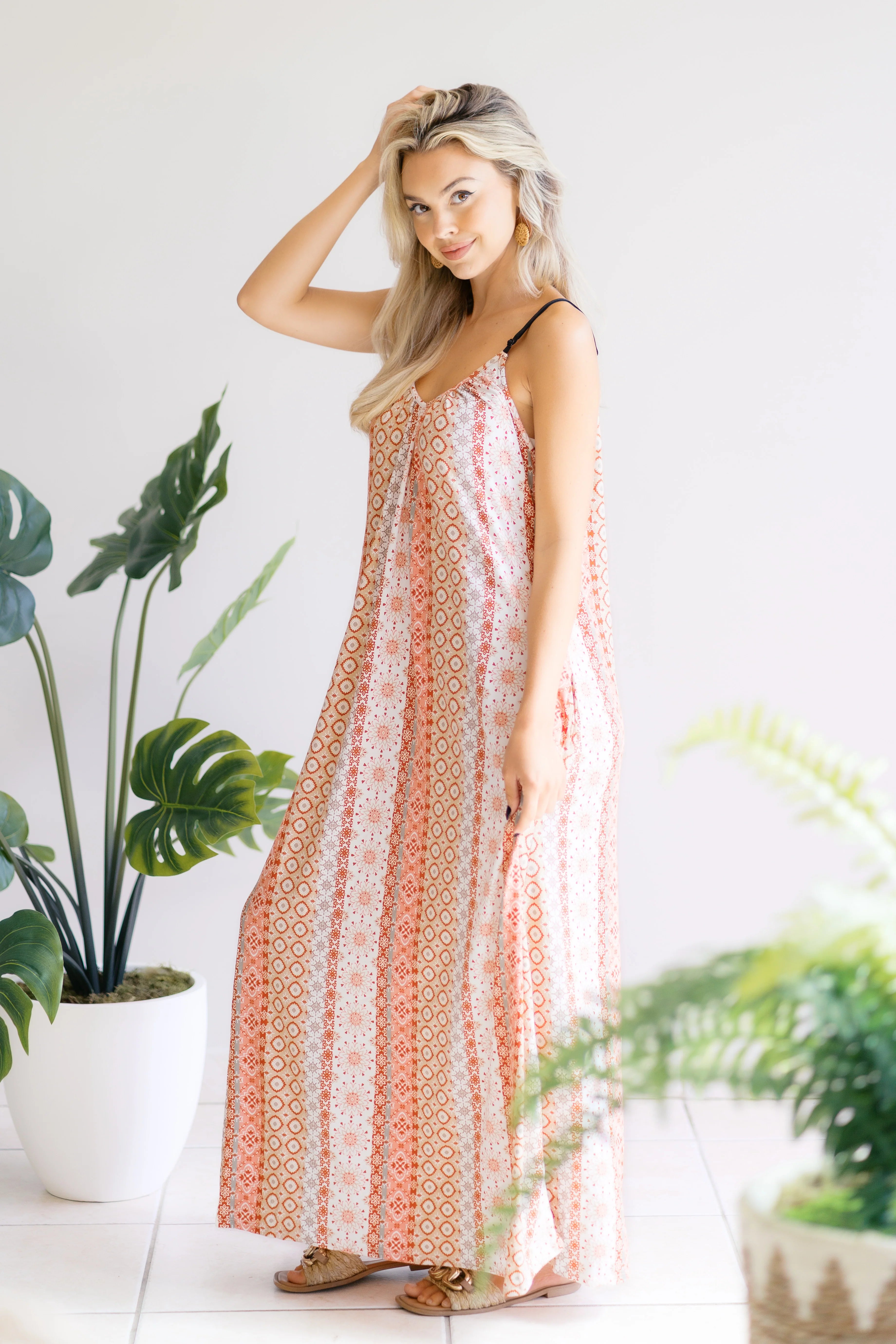 Blonde woman in sleeveless patterned maxi dress standing indoors near large potted tropical plants
