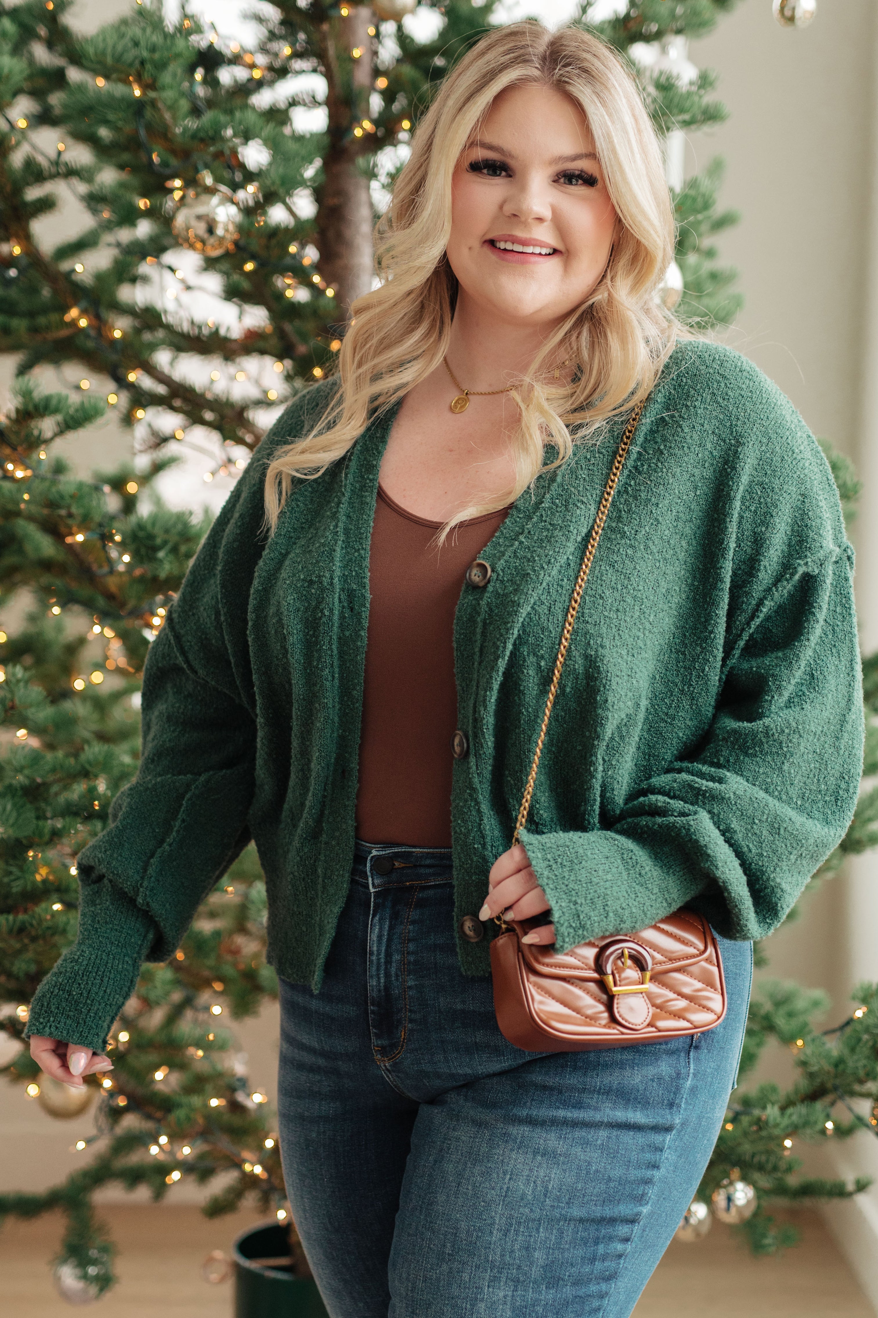 Plus size woman wearing a cozy moss green button-up cardigan over a brown top and blue jeans by decorated Christmas tree