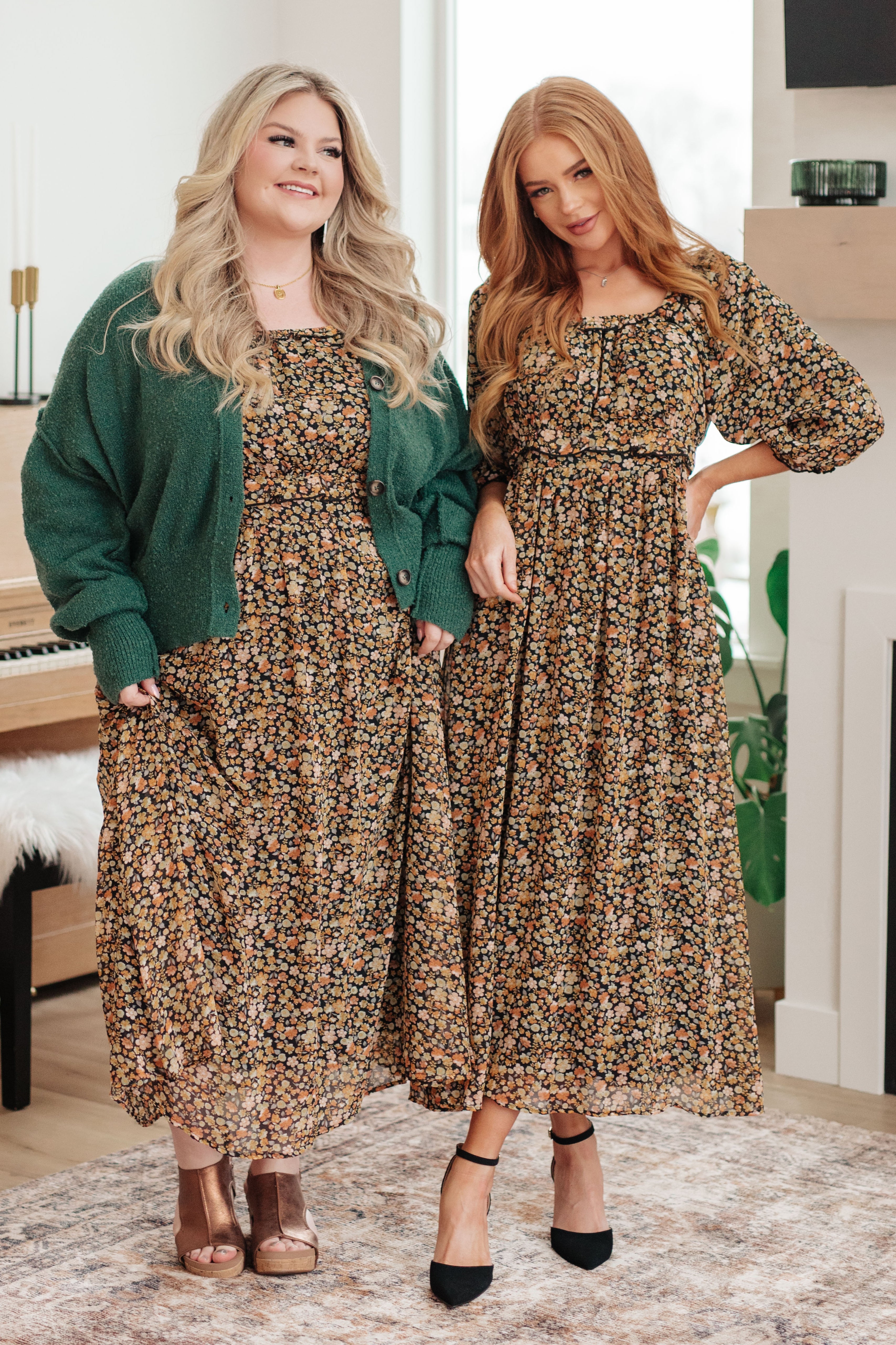 Two women wearing floral maxi dresses, one paired with a dark green button-up cardigan indoors by piano and plants