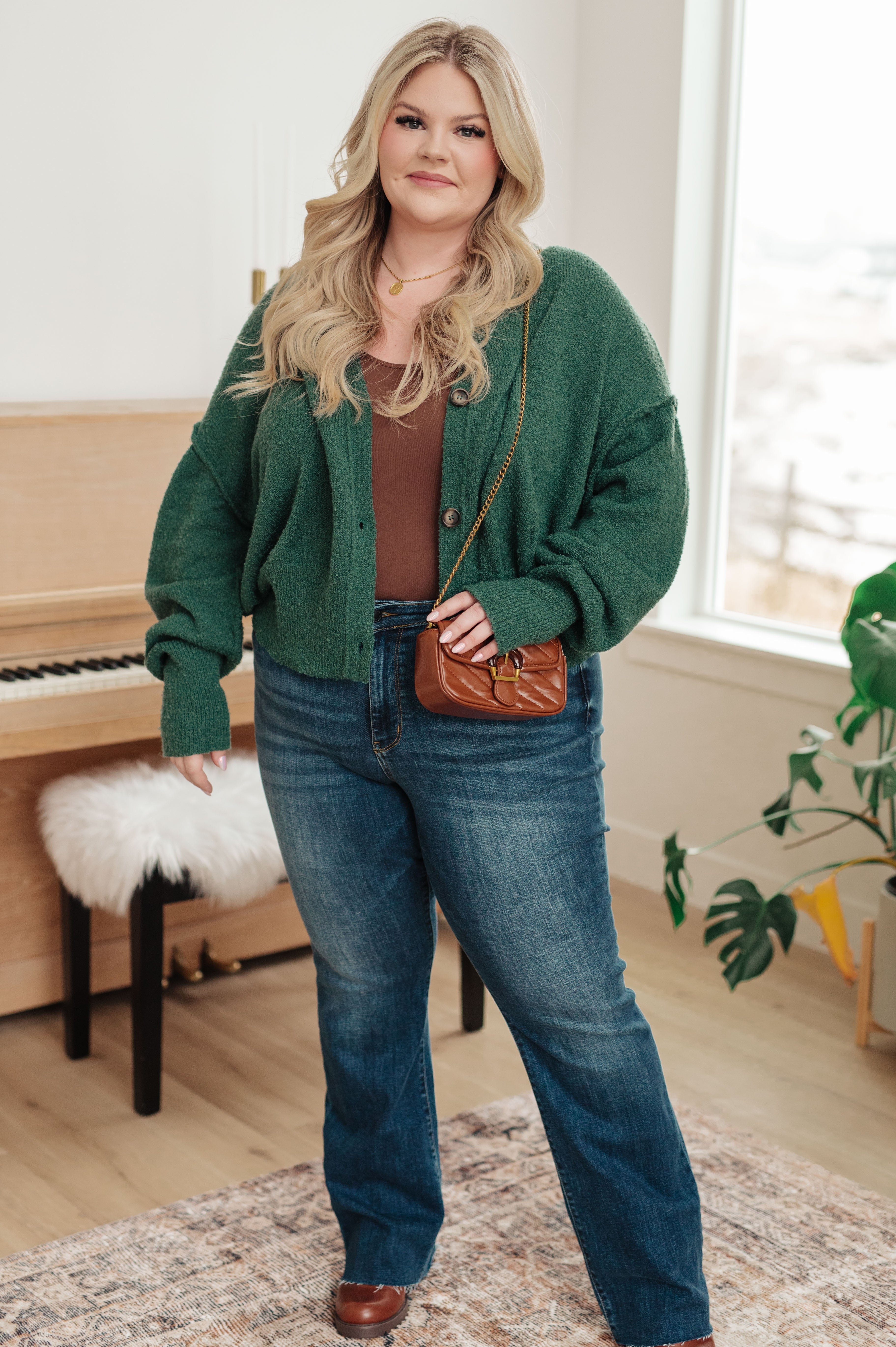 Plus size woman wearing dark green button-down cardigan with blue jeans and brown crossbody bag indoors near piano
