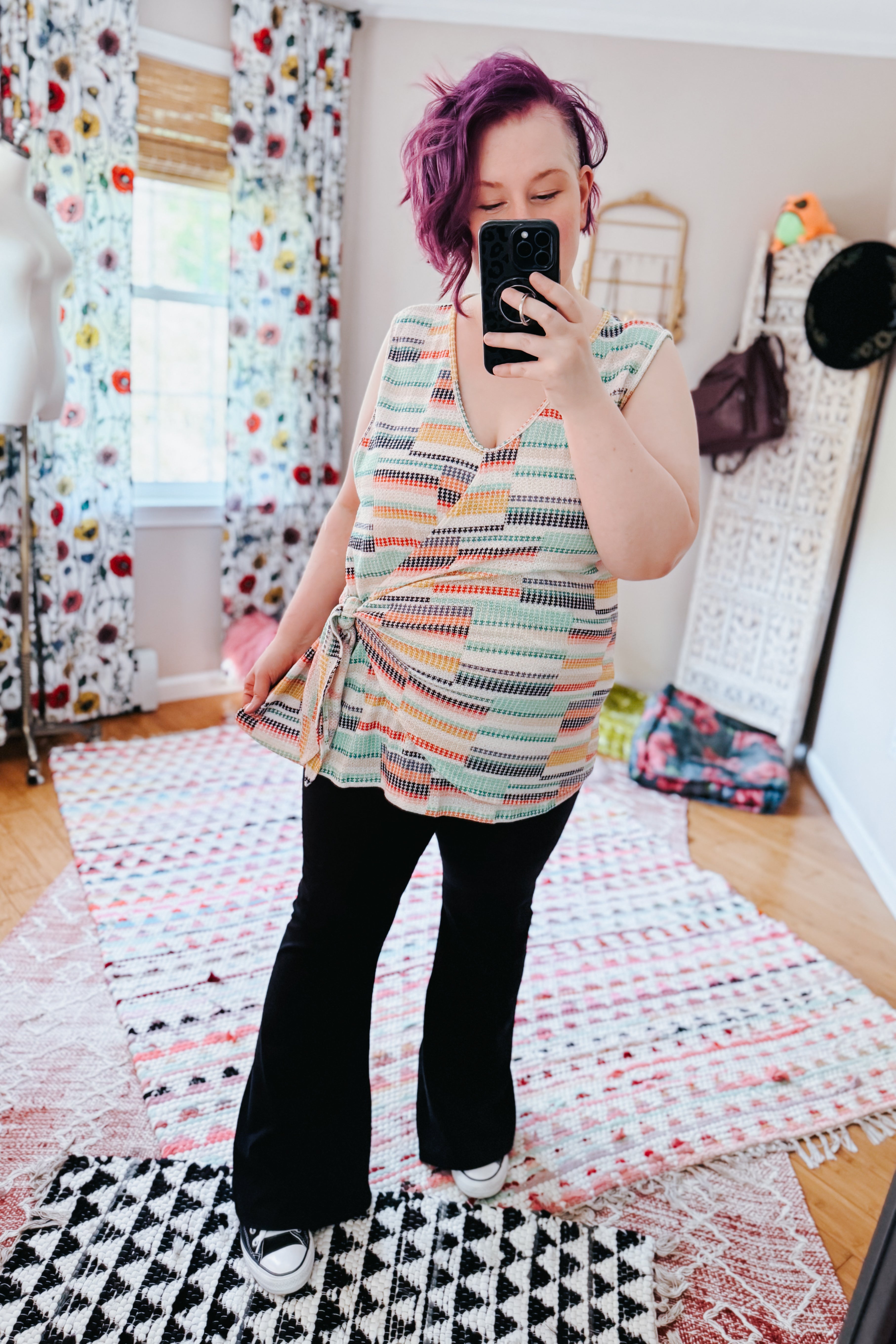 Woman with purple hair wearing a colorful geometric wrap tank and black pants in a free-spirited eclectic room
