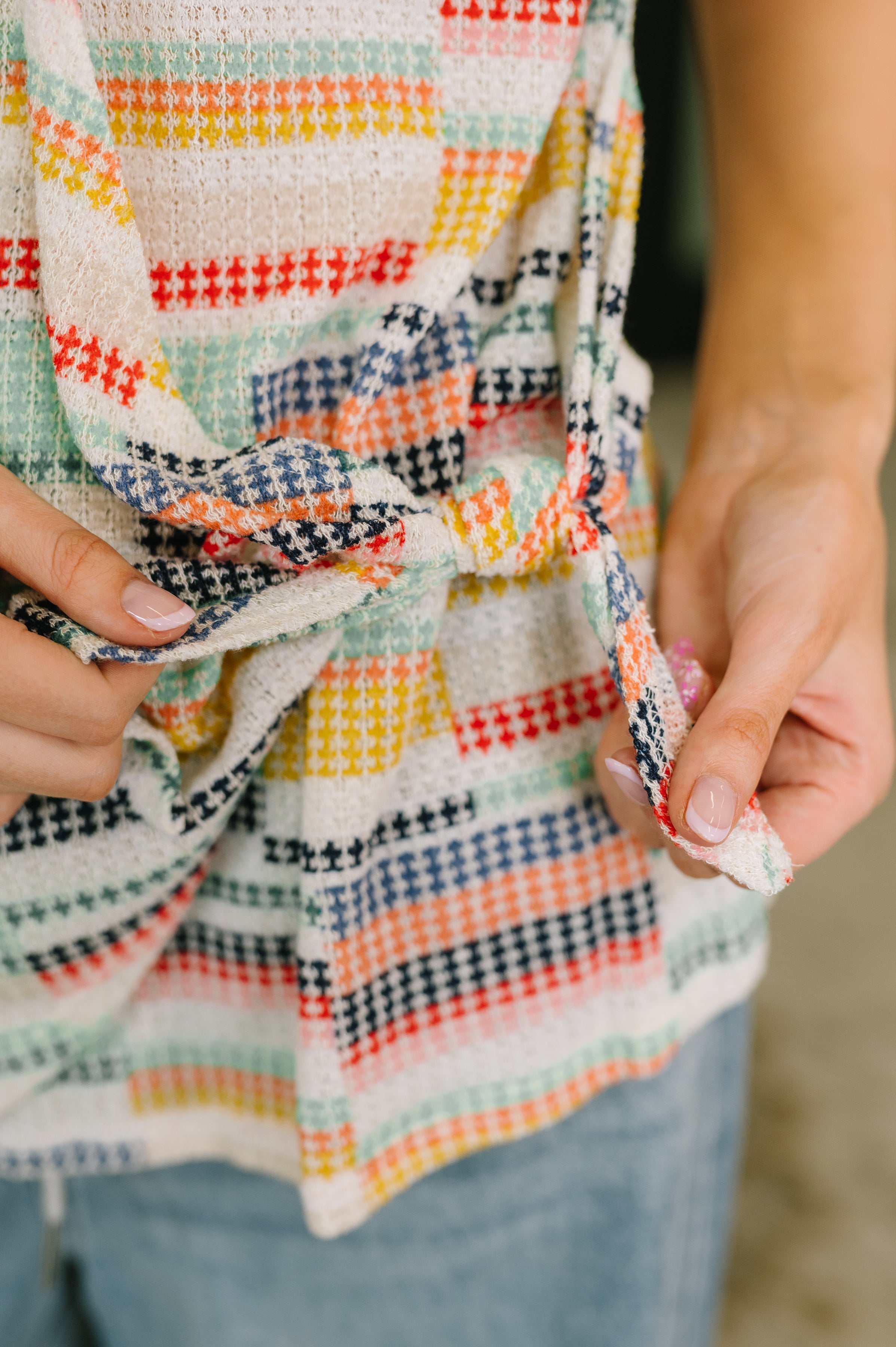 Close-up of person tying a colorful patterned top with textured fabric, paired with light blue jeans and natural nails.