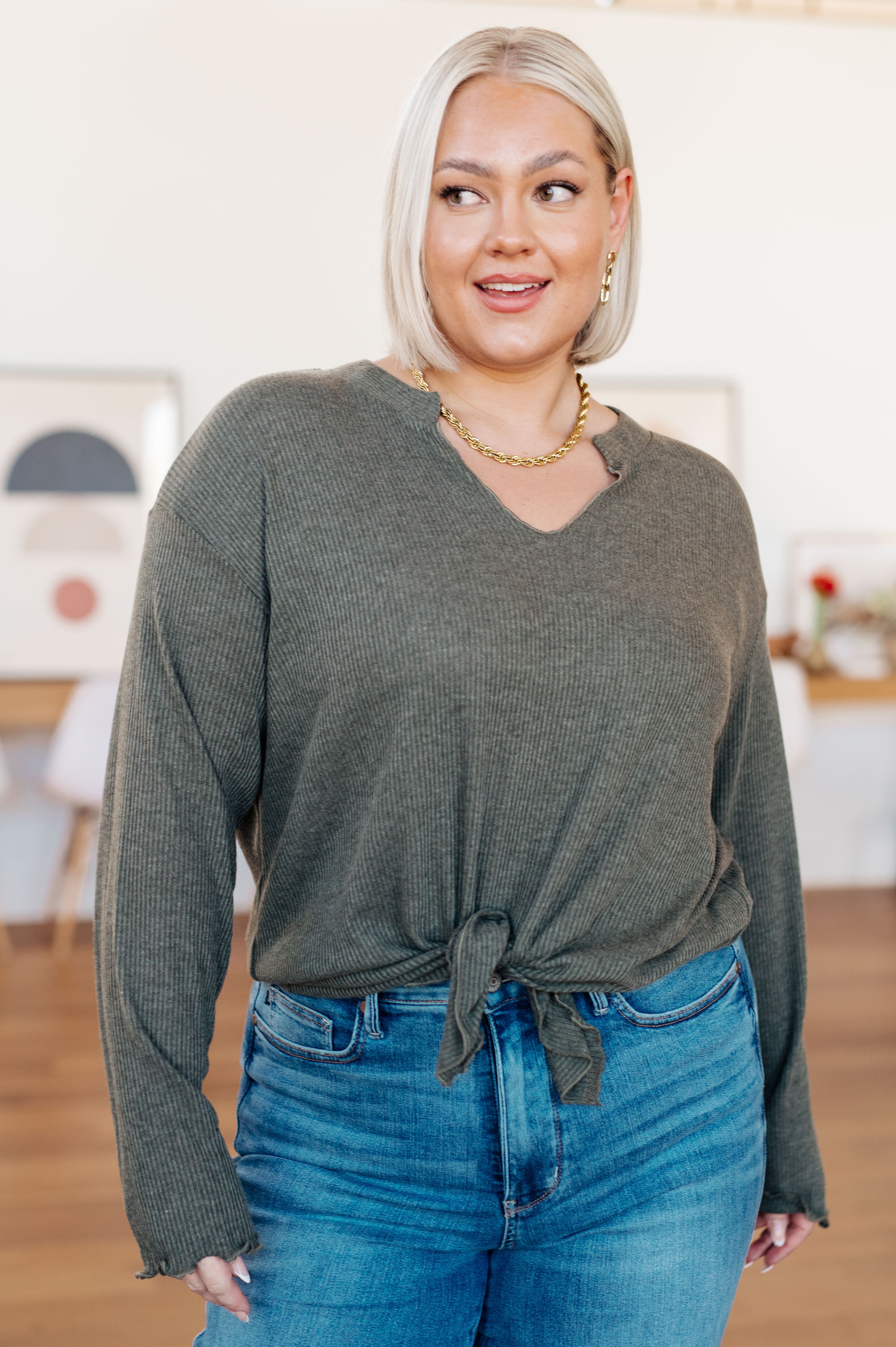 Woman wearing a grey ribbed knit top with a tie front and V neckline paired with blue jeans in a bright room