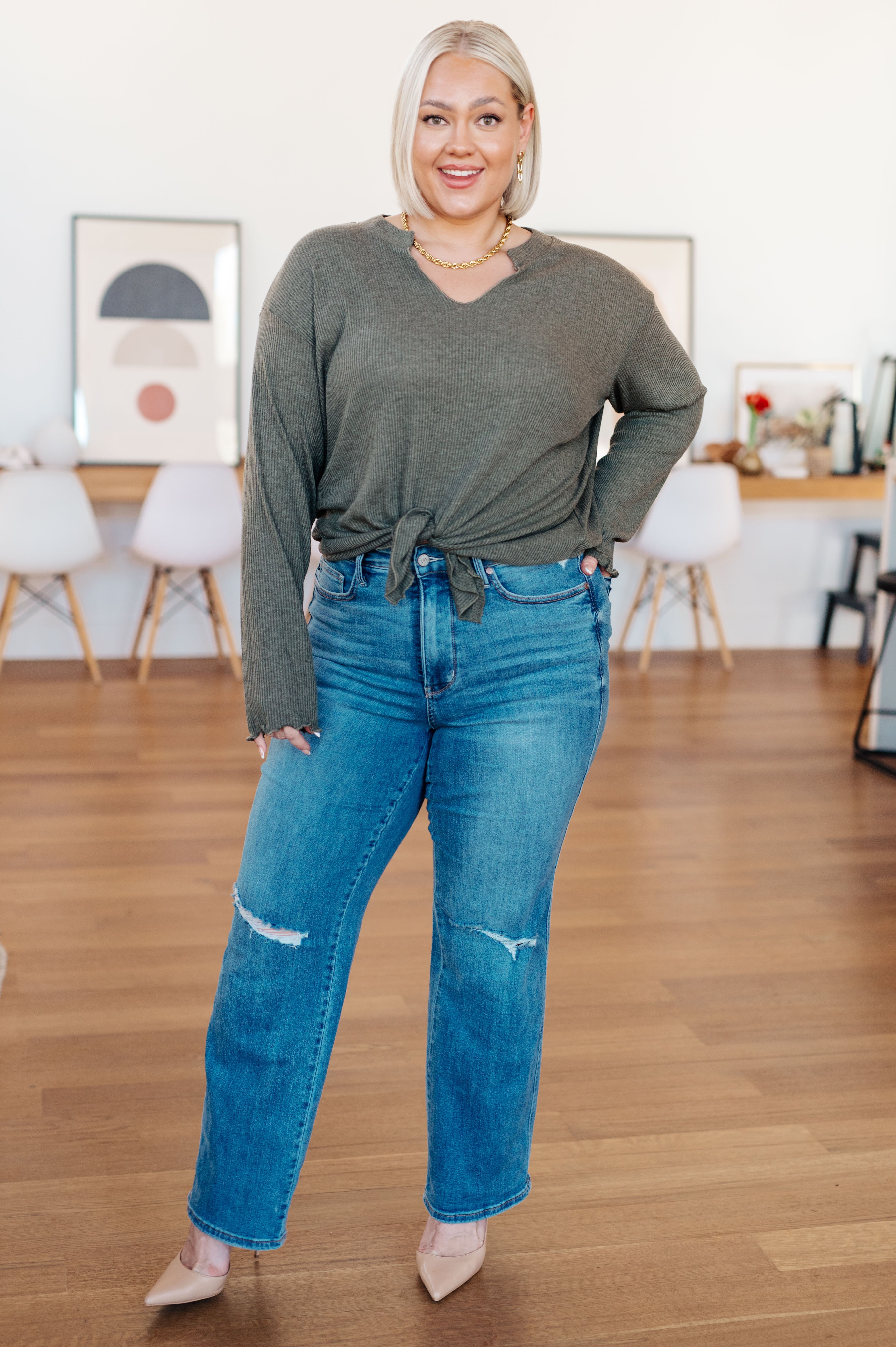 Smiling woman in a grey ribbed knit top with a V neckline and tie front styled with distressed blue jeans indoors
