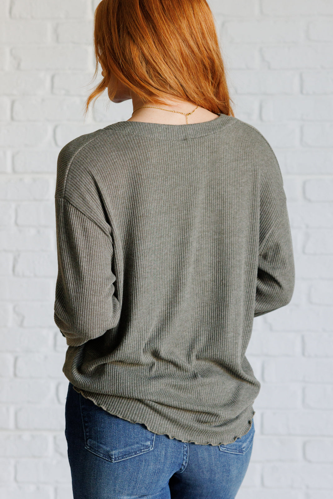 Back view of woman wearing a grey ribbed knit top with long sleeves and lettuce hem paired with blue jeans against a white brick wall