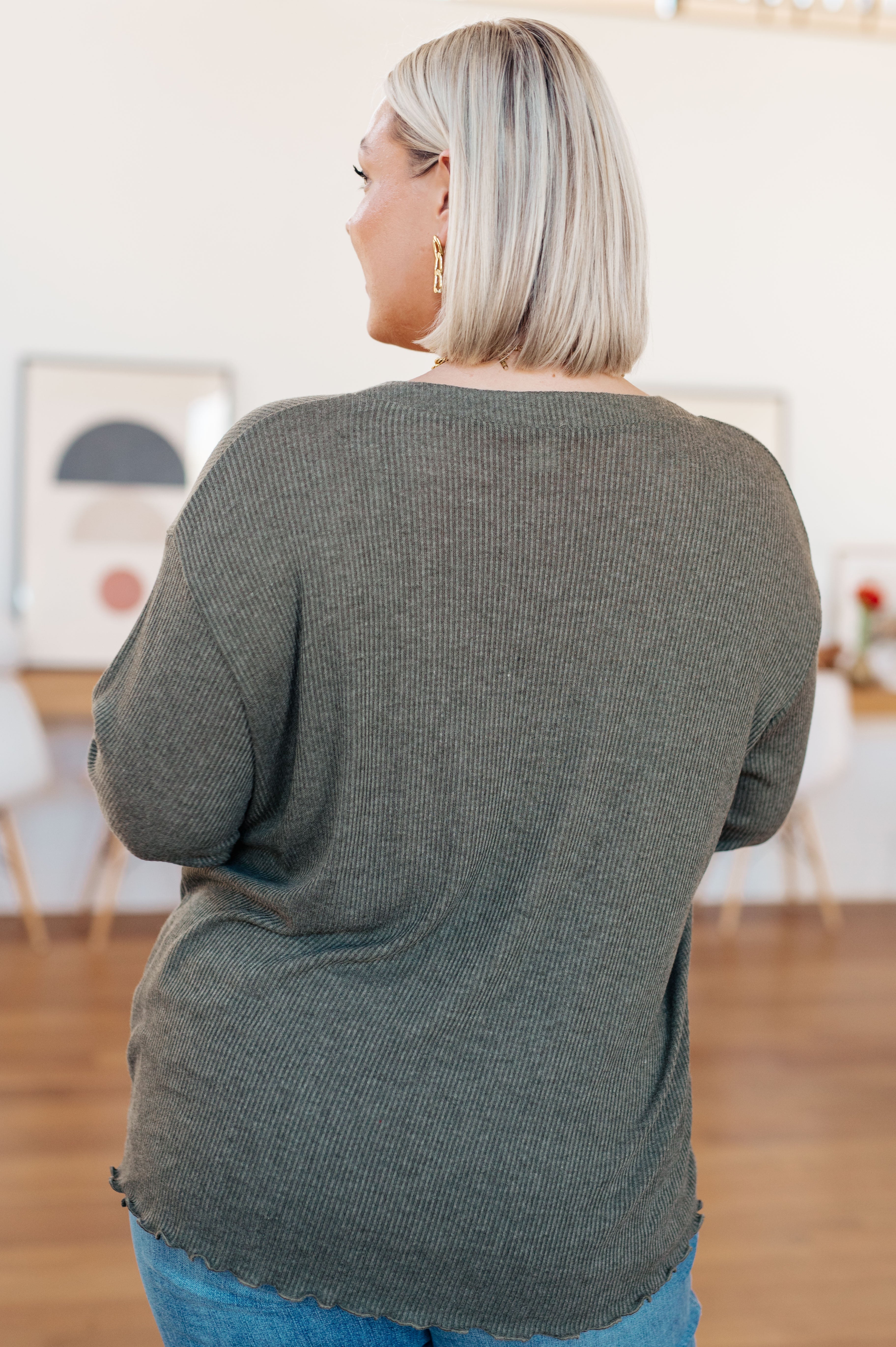 Back view of a woman wearing a grey ribbed knit top with a lettuce hem and blue jeans in a warmly styled indoor room