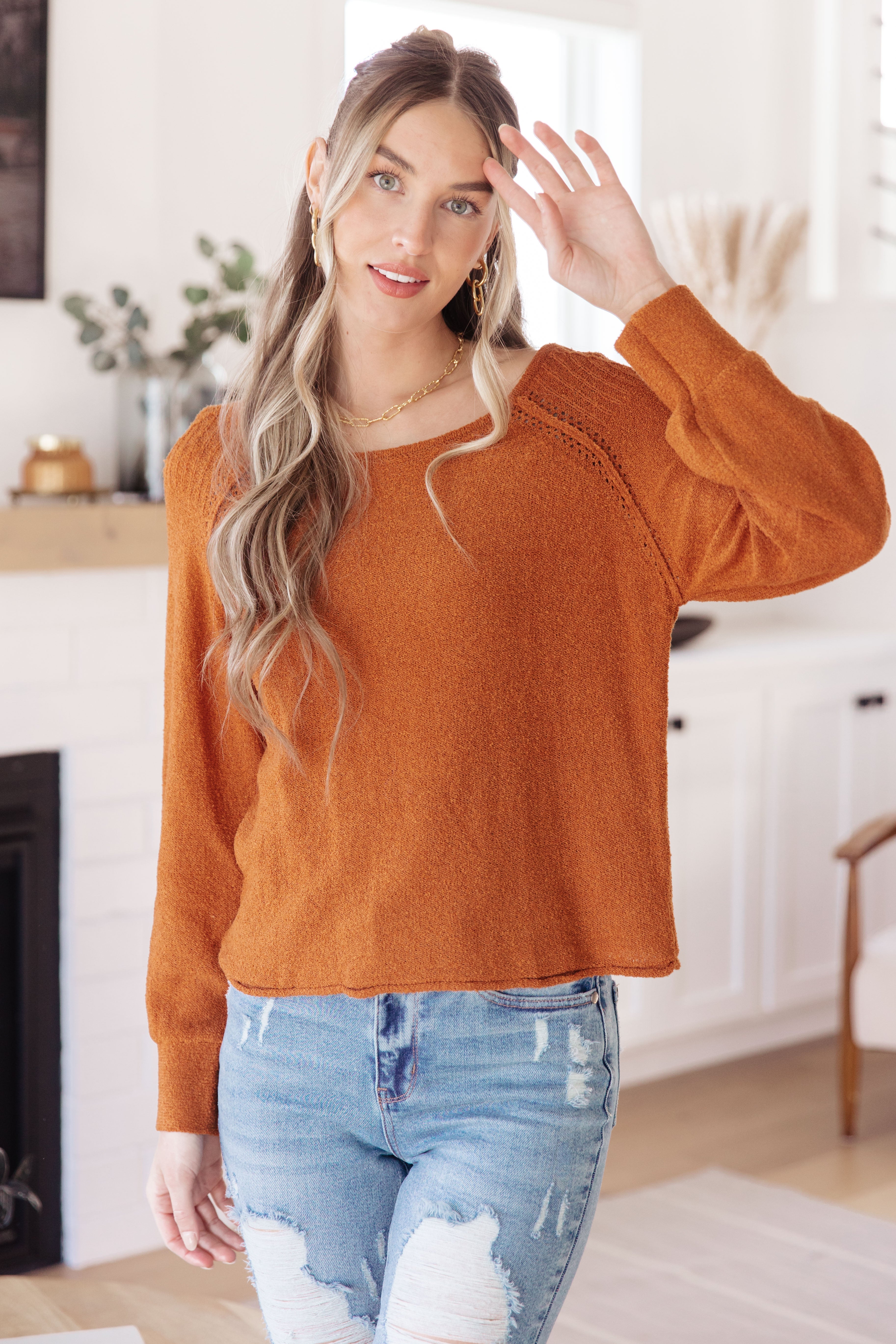 Woman with long wavy hair wearing a textured burnt orange pullover sweater and ripped light wash jeans indoors