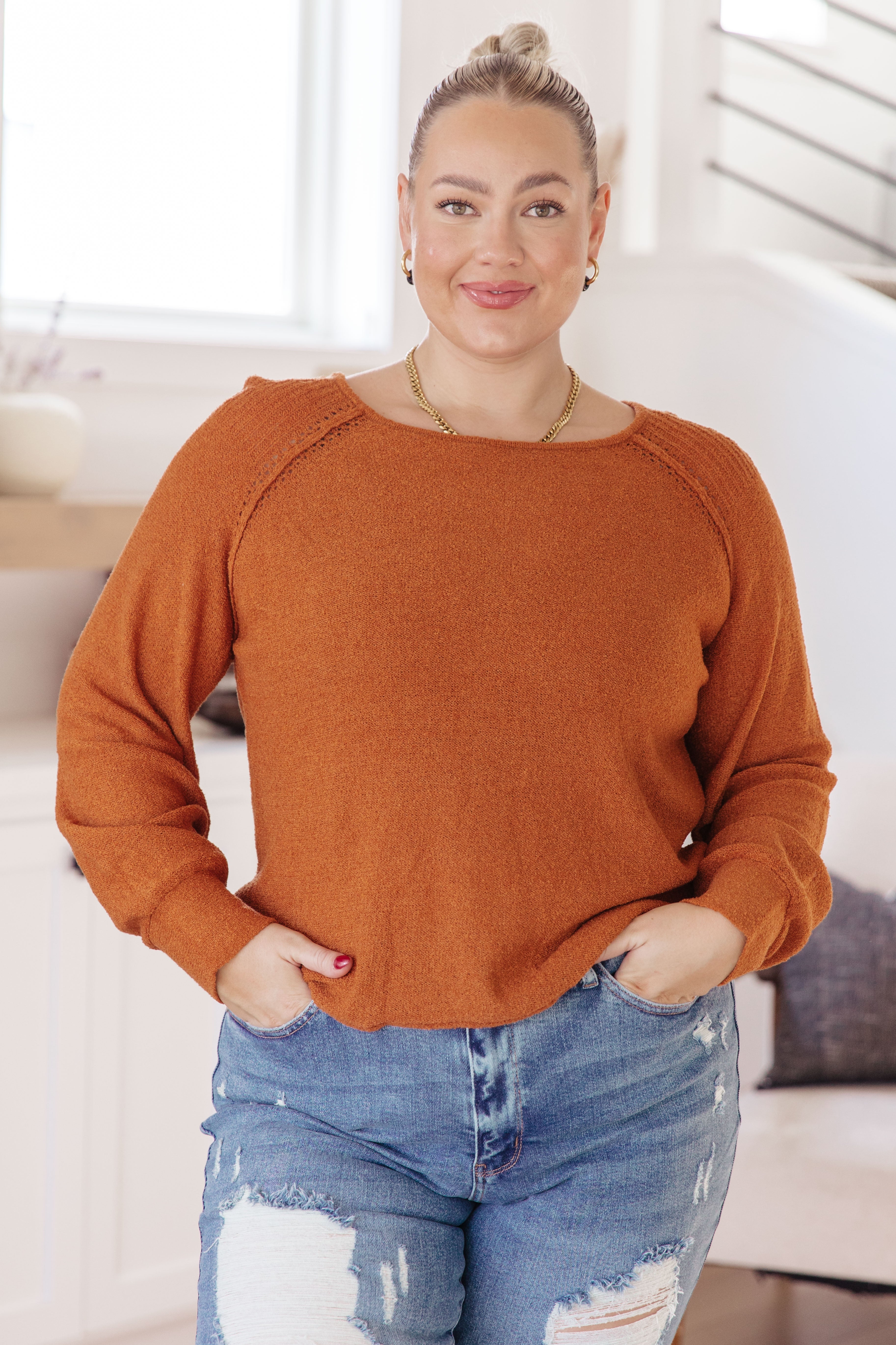 Smiling woman with blonde hair in a bun wearing a burnt orange pullover sweater and ripped denim jeans indoors