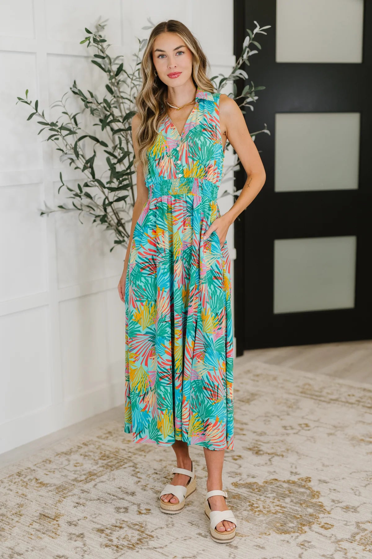 Woman wearing a sleeveless mint dress with colorful tropical print, standing indoors with one hand in pocket