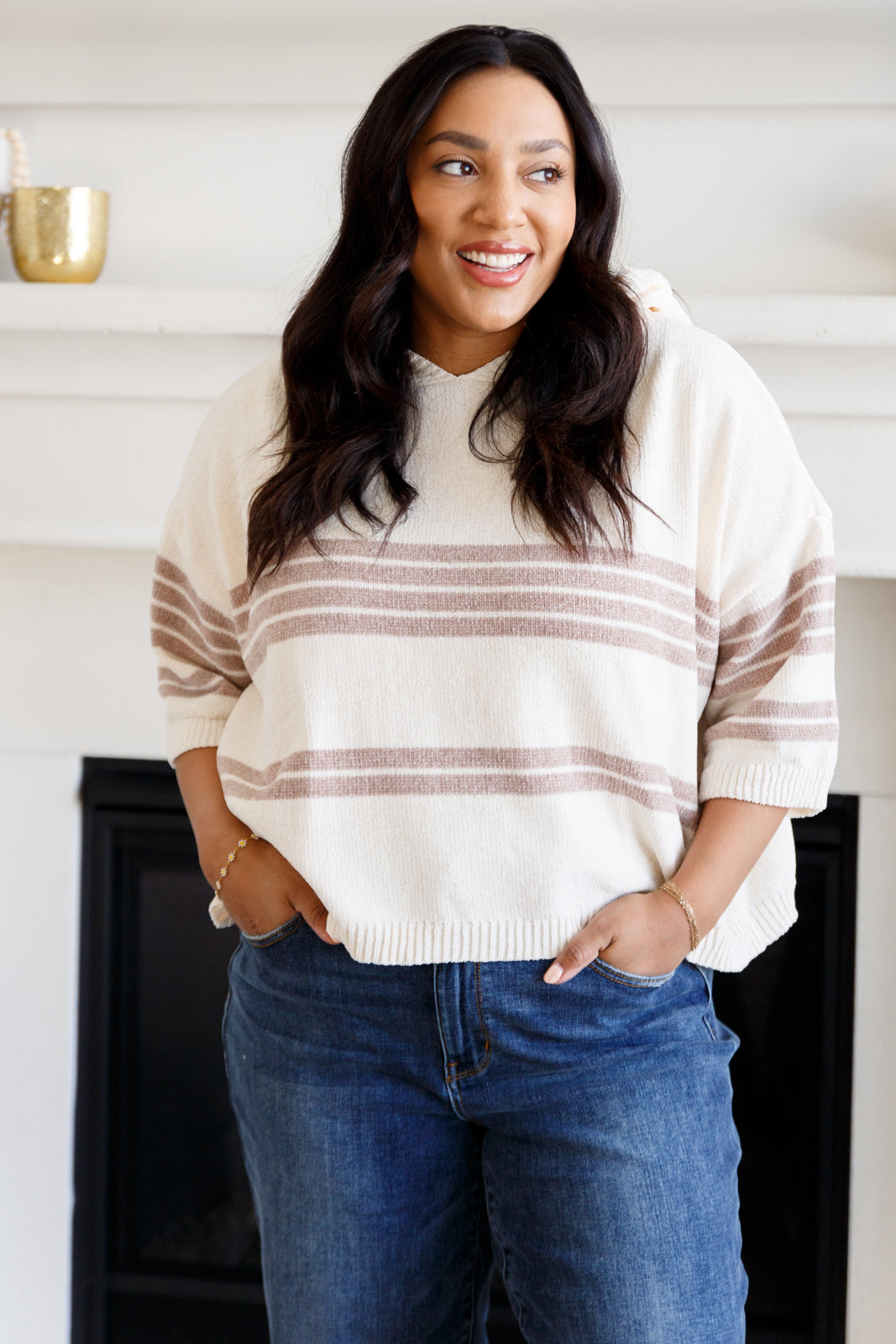 Woman smiling indoors wearing cream hoodie with light taupe stripes and relaxed fit blue jeans with hands in pockets