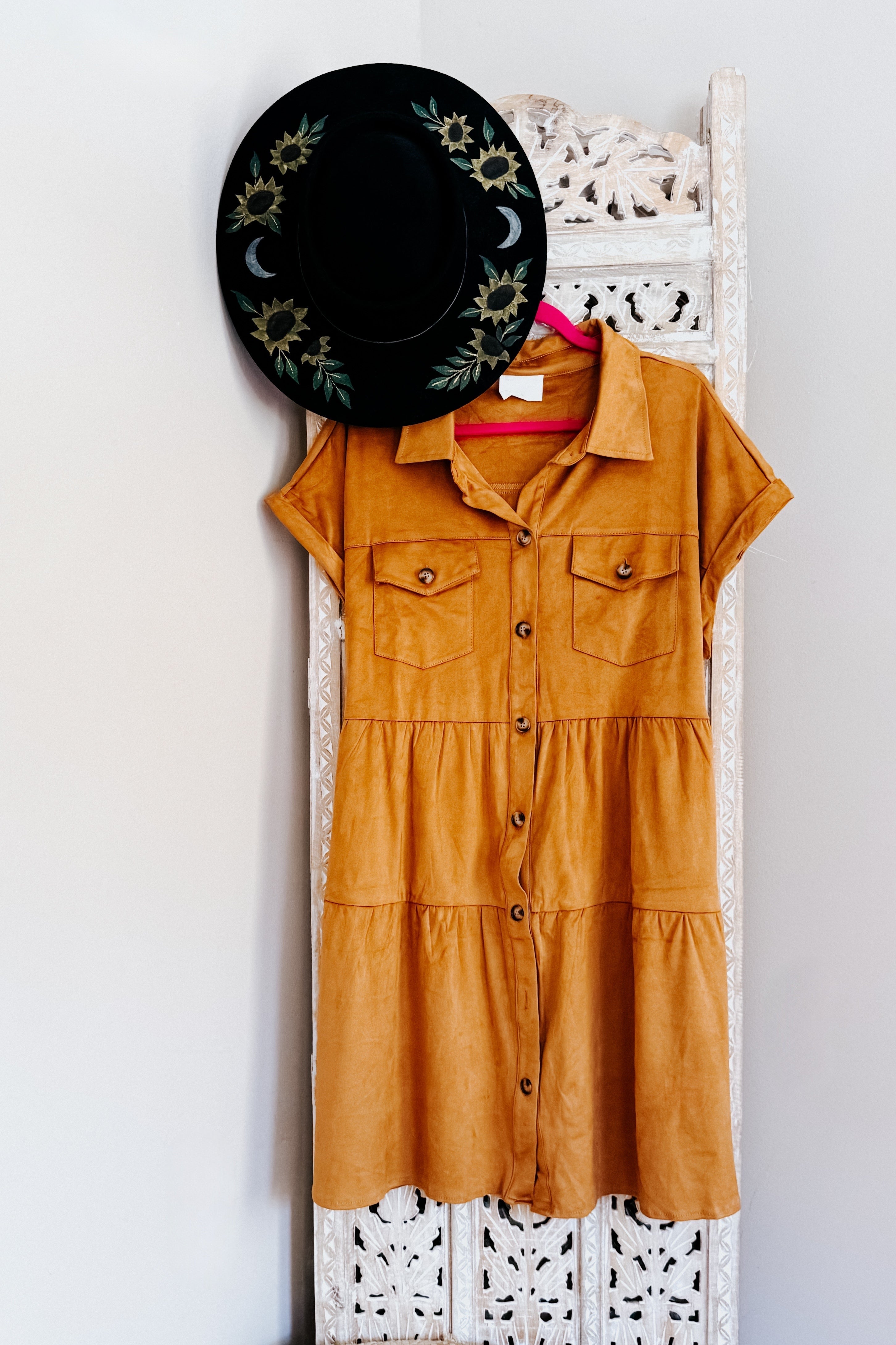 Tan faux suede short-sleeve shirt dress with button front and chest pockets hanging on carved wooden panel with black hat