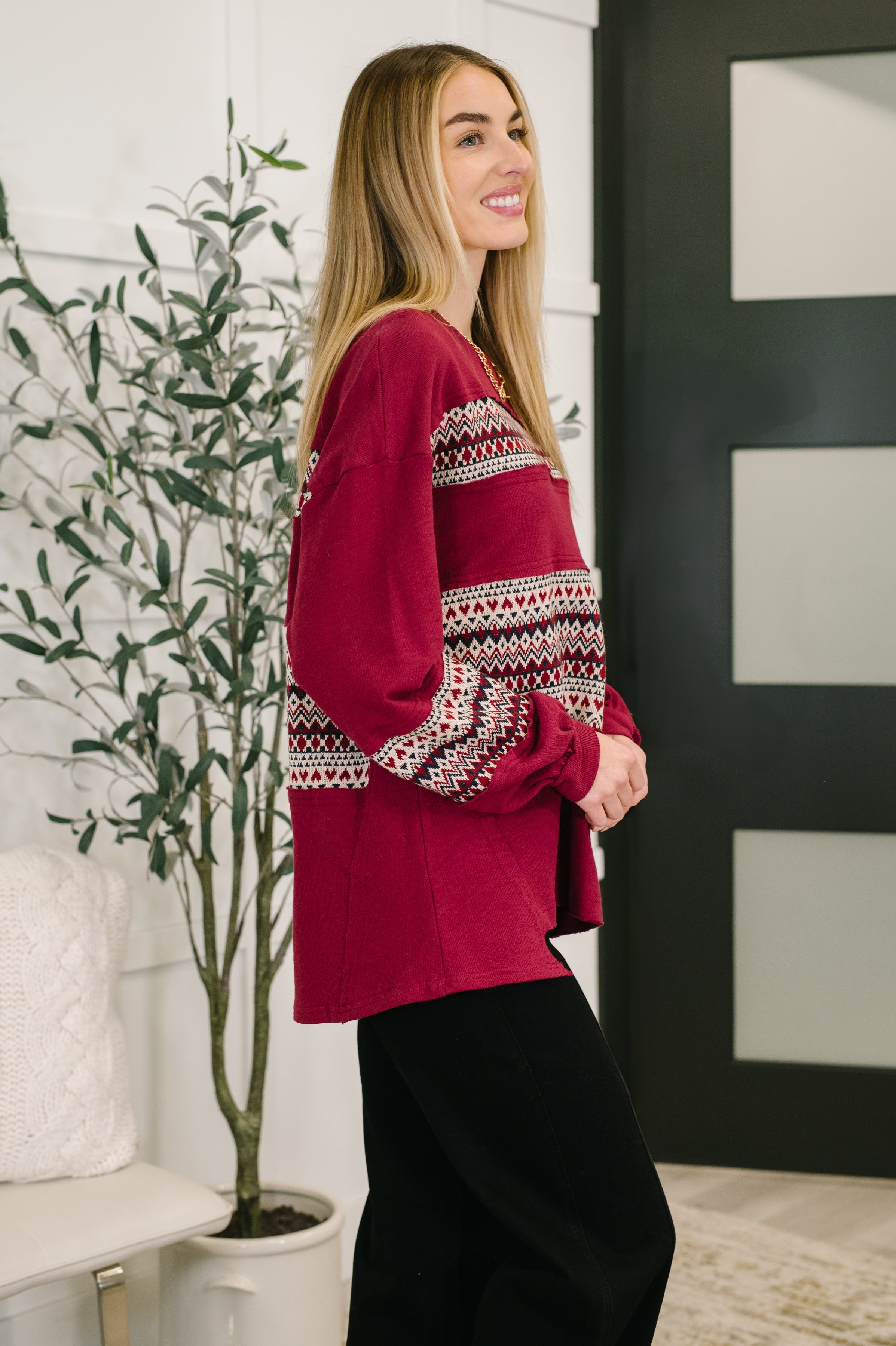 Woman wearing a long sleeve patterned red sweater, a gold necklace and black pants standing in an indoor setting.