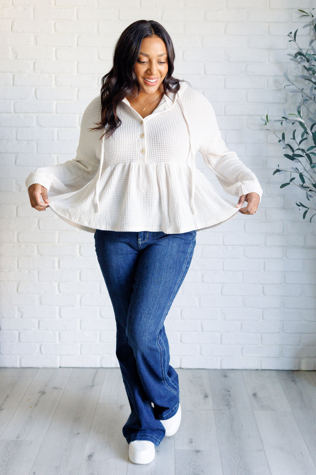 Smiling woman wearing cream waffle knit hoodie with buttons and flared hem paired with dark blue jeans and white shoes indoors