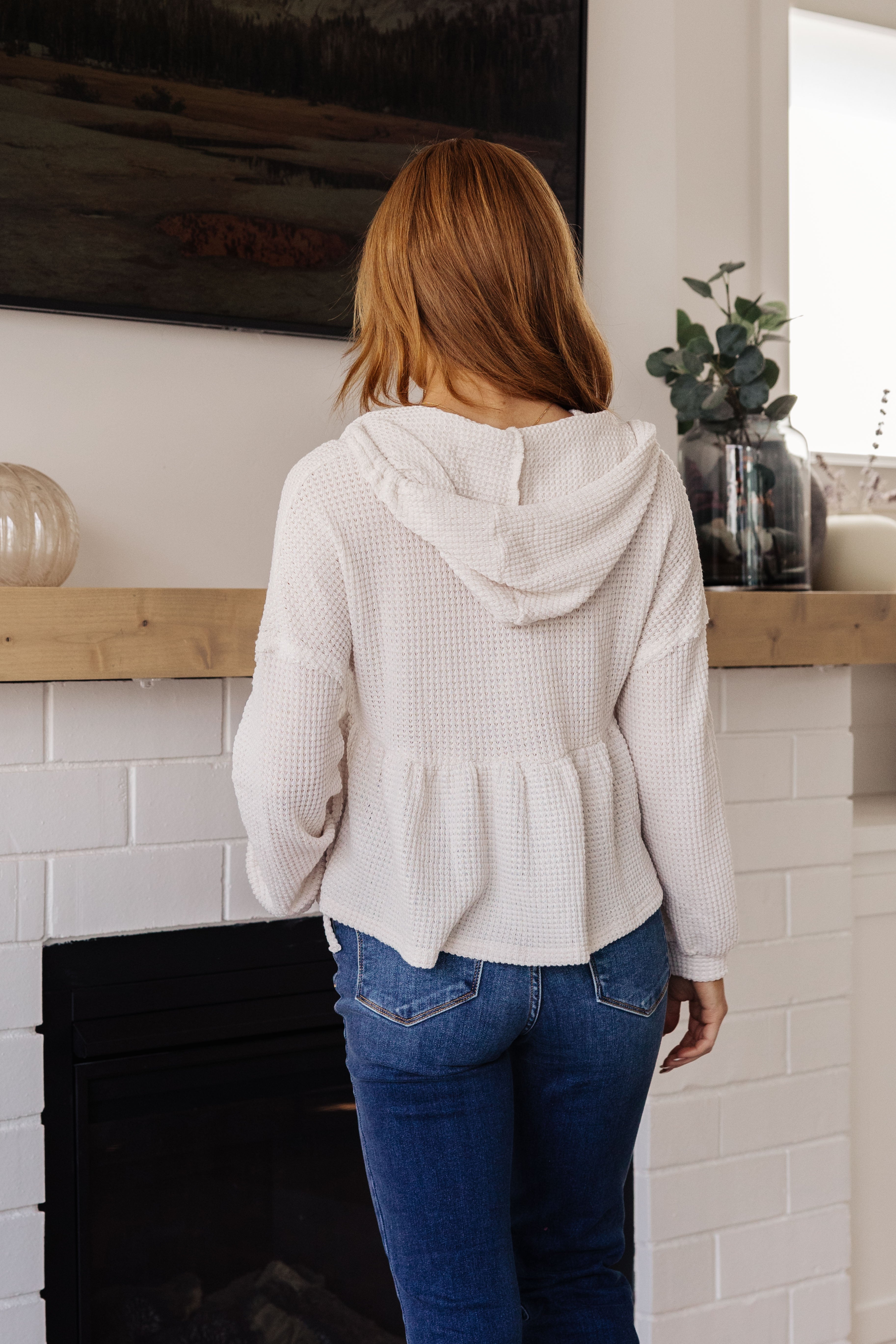 Redhead woman indoors wearing off-white waffle knit hoodie with gathered peplum hem and blue jeans standing by fireplace