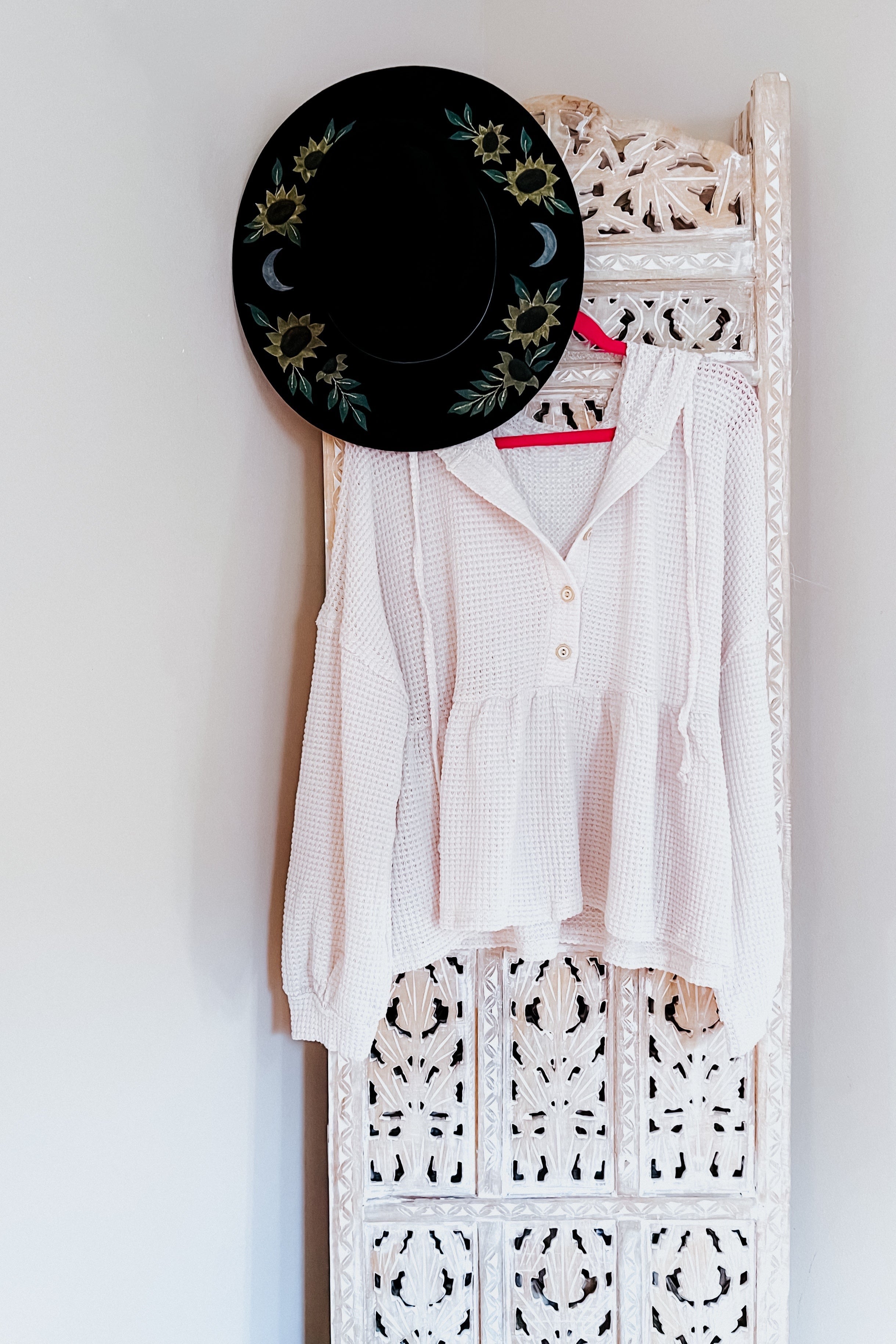 Cream waffle knit hoodie with buttons and peplum hem hanging on white carved wooden panel with black floral hat