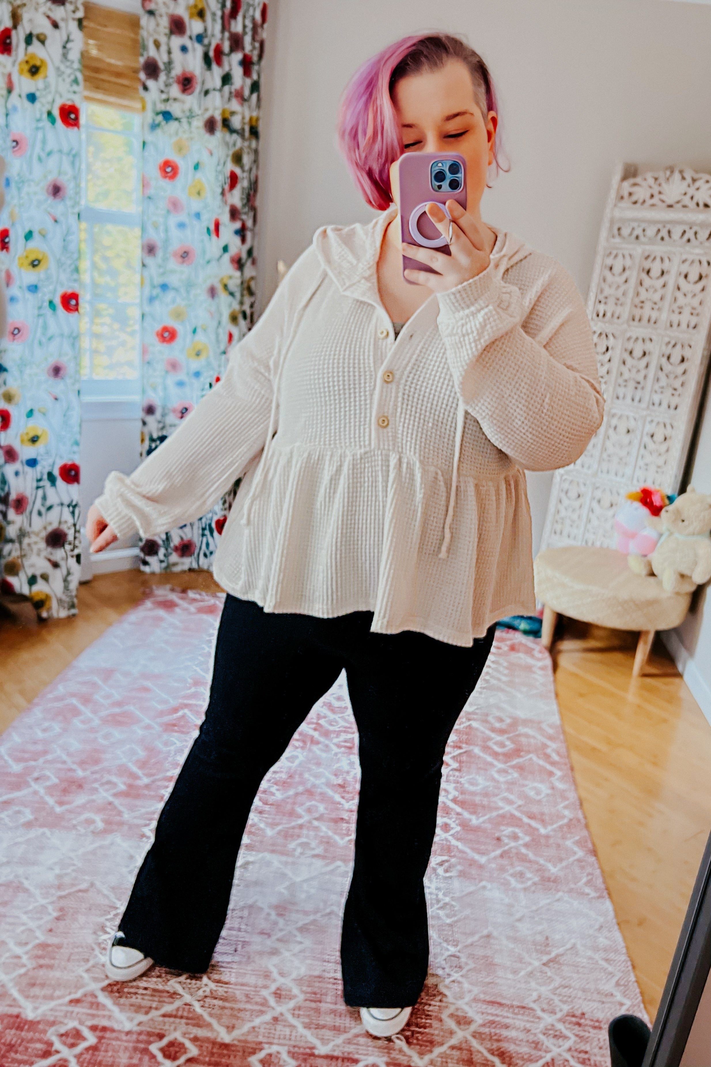 Woman with short pink hair wearing off-white waffle knit hoodie with button front and peplum hem taking mirror selfie indoors