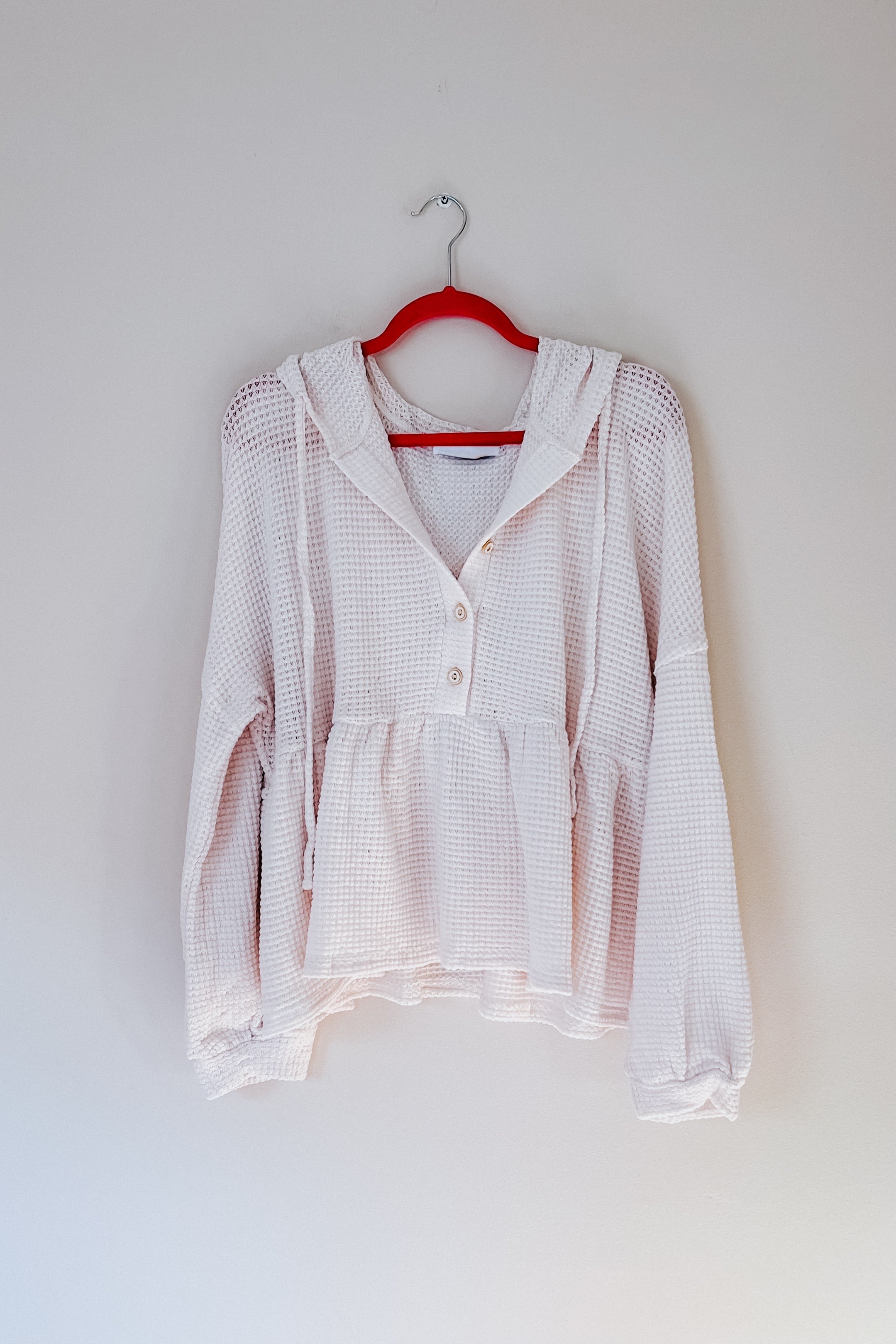 Light pink waffle knit hoodie with buttons and gathered waist hanging on red hanger against plain wall