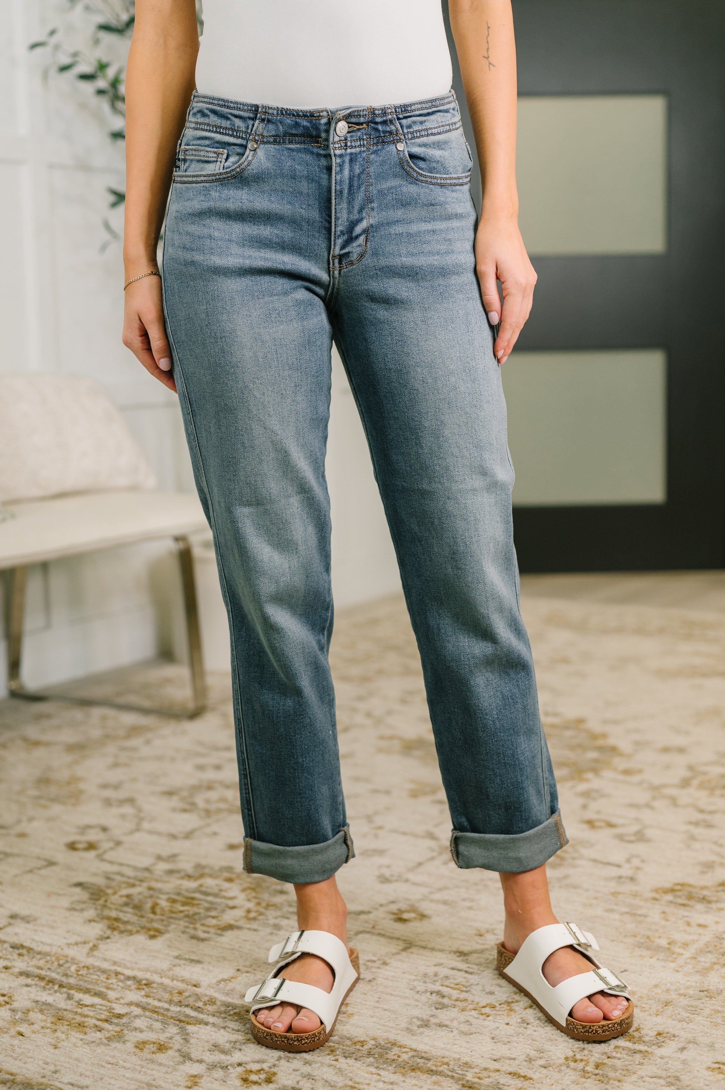 Blue mid rise boyfriend jeans with rolled cuffs worn with white sandals indoors on carpeted floor