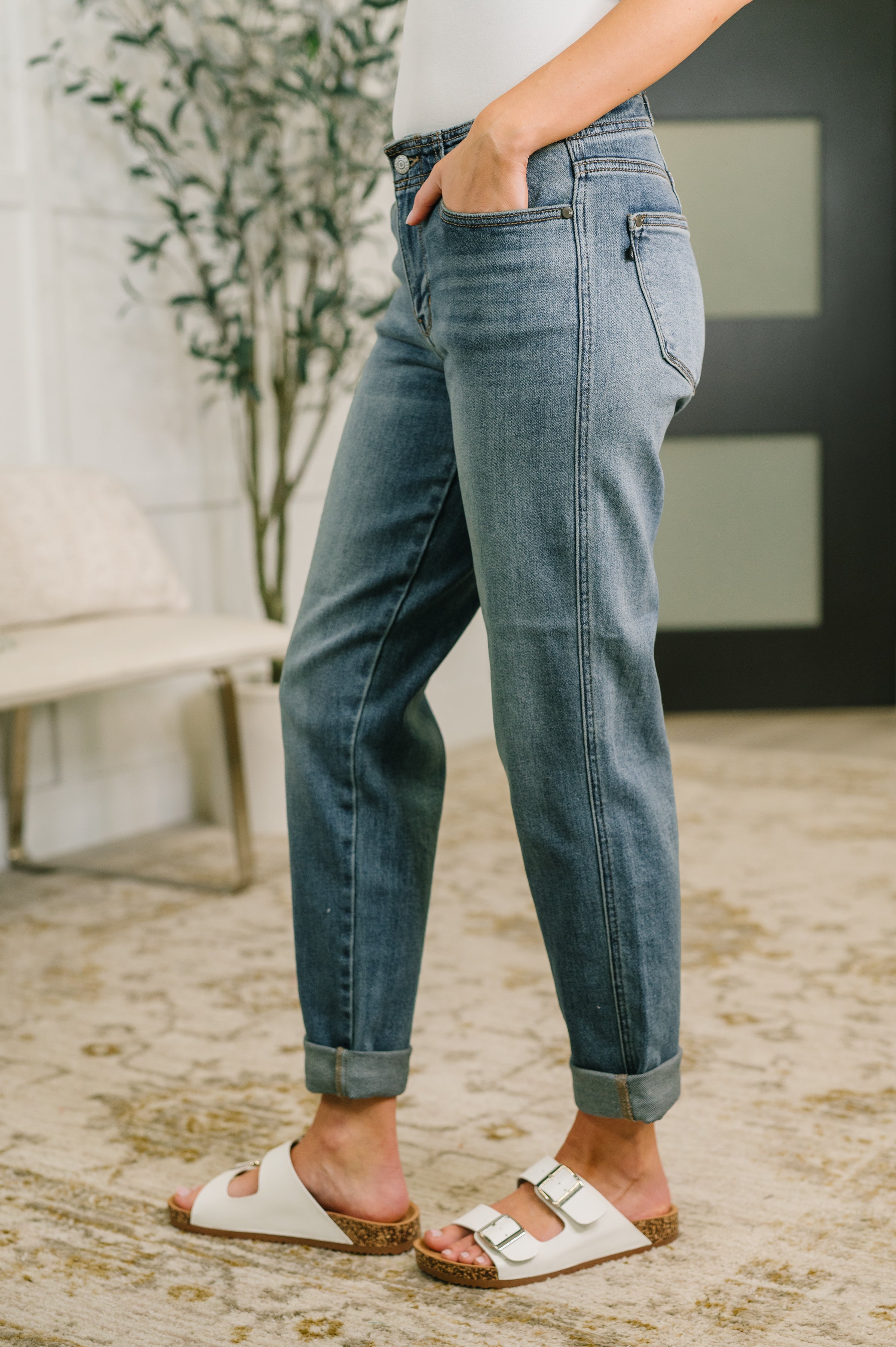 Woman wearing mid rise blue denim boyfriend jeans with rolled cuffs and white buckle sandals indoors