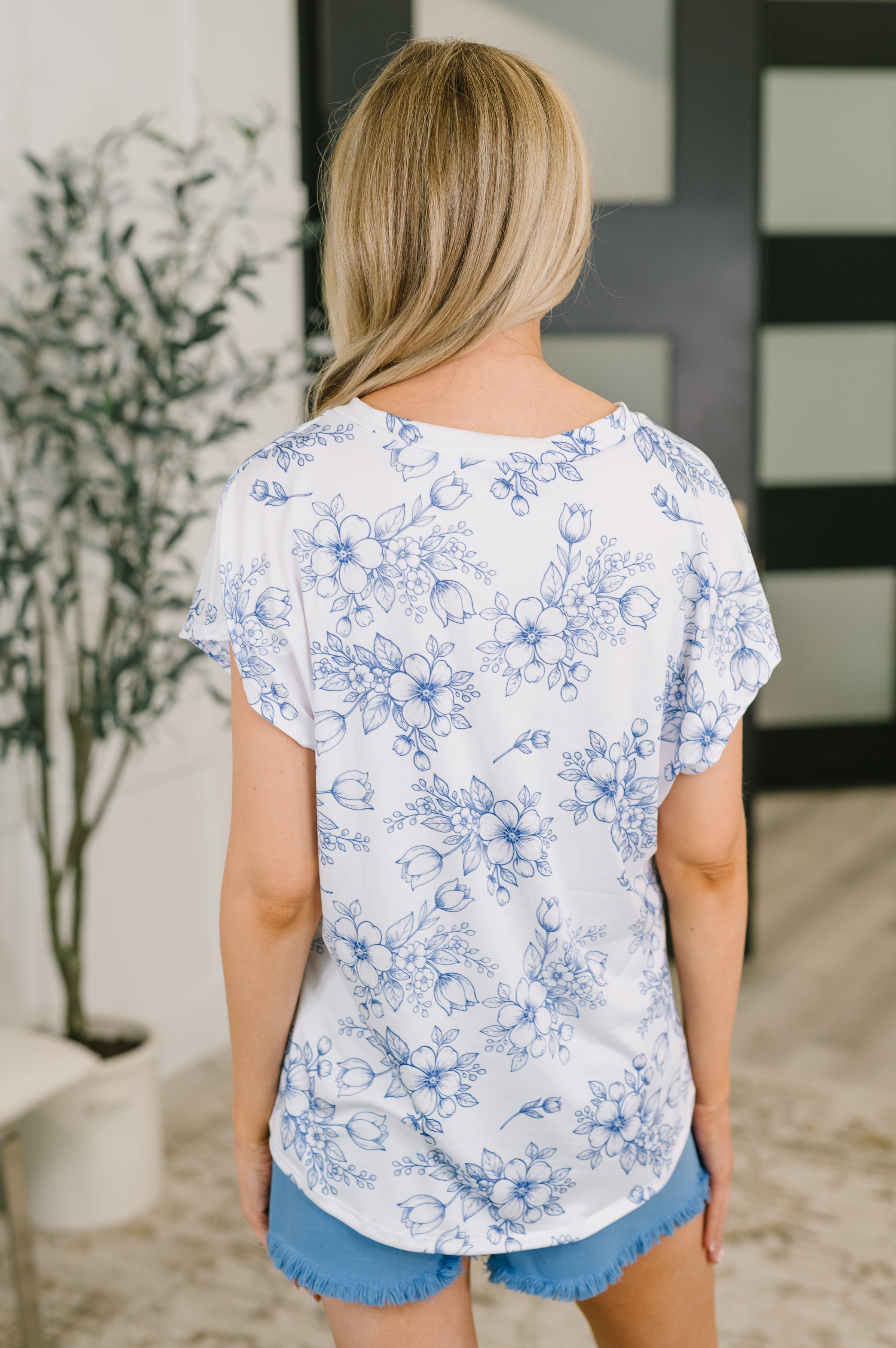 Back view of woman wearing a cream floral top with blue sketch-style flowers paired with blue frayed shorts indoors