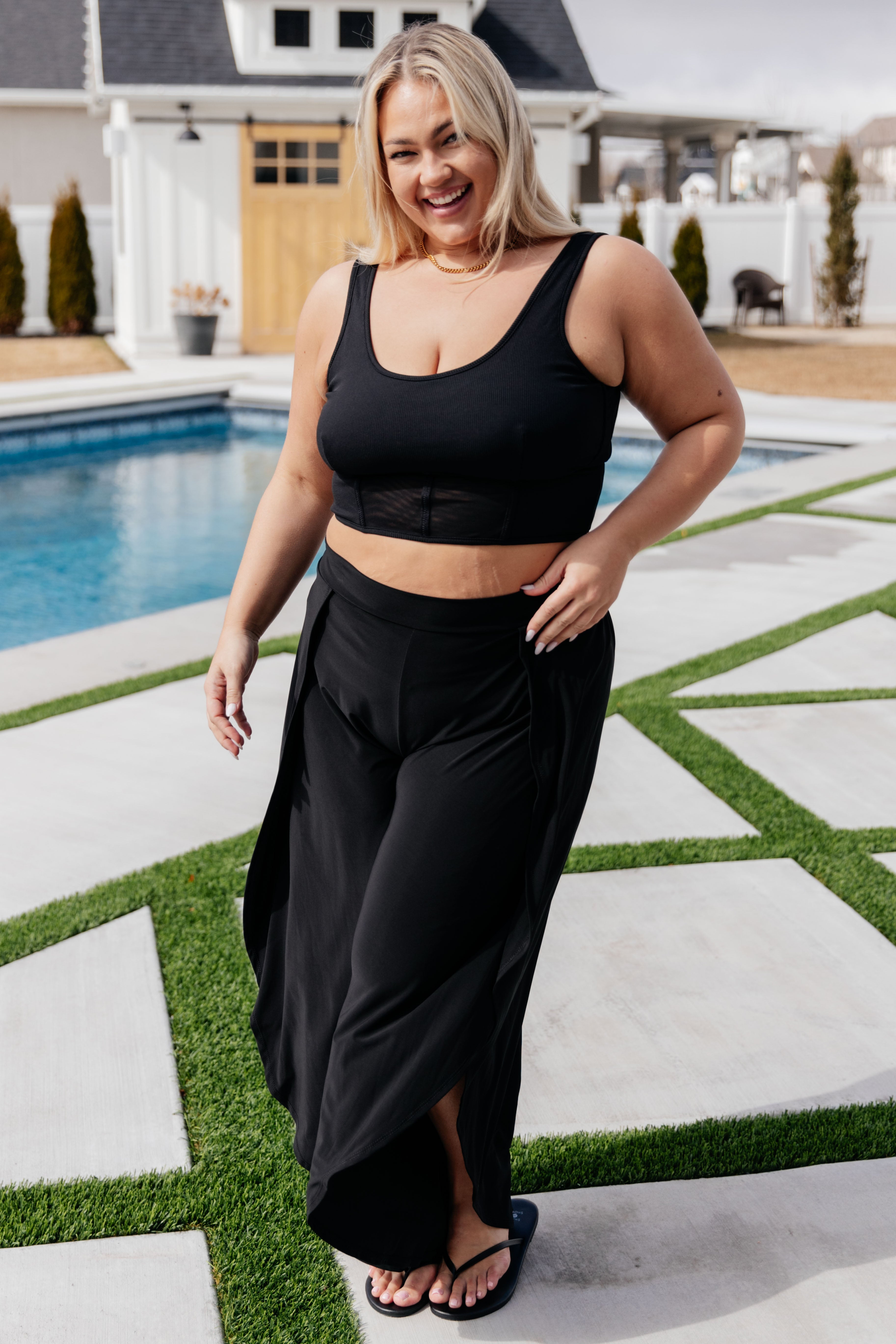 Smiling woman wearing a black scoop neck crop top and wide-leg pants walking near a pool outdoors