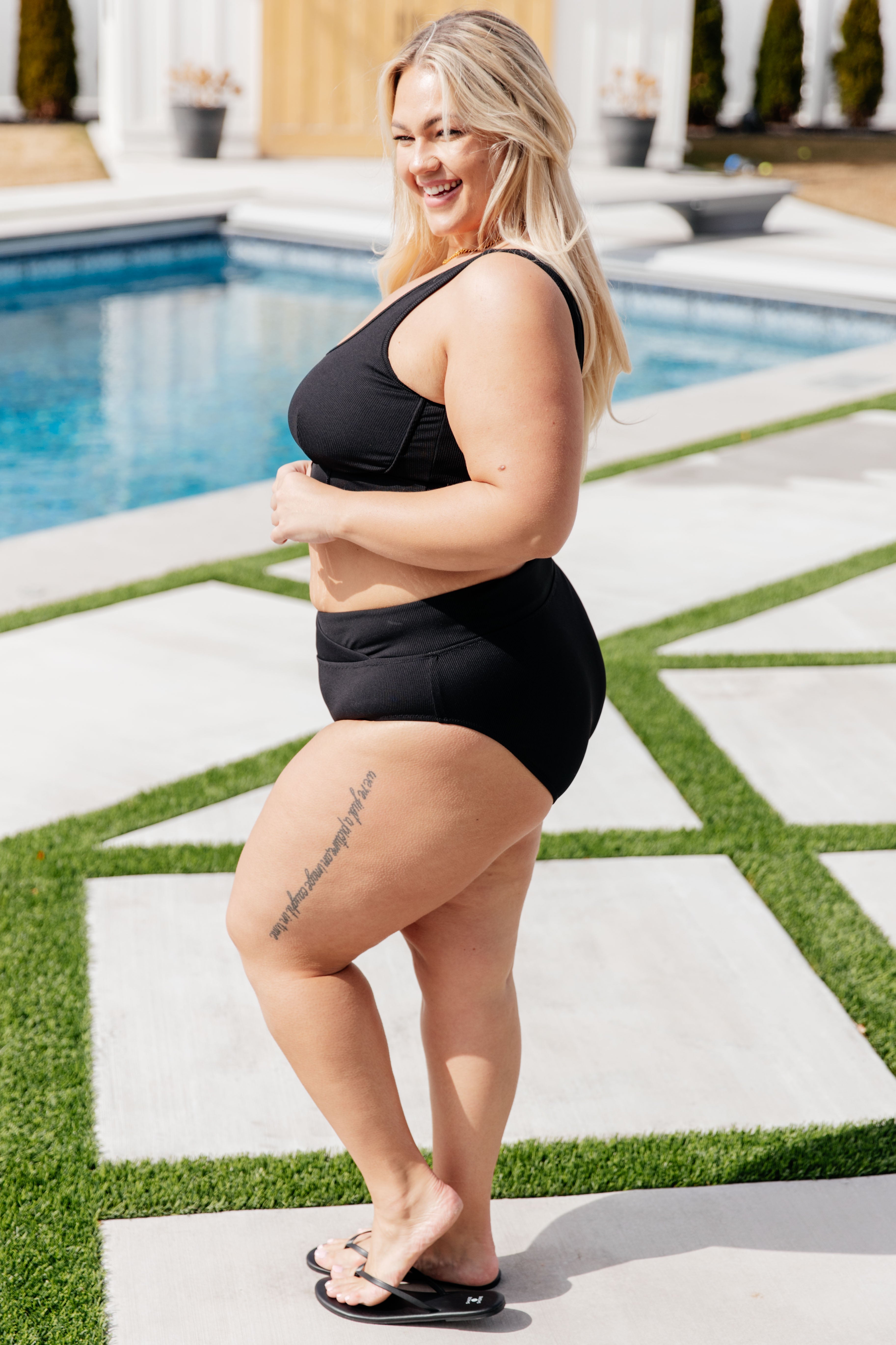 Smiling woman wearing a black two-piece swimsuit and black flip-flops standing near an outdoor pool on a sunny day