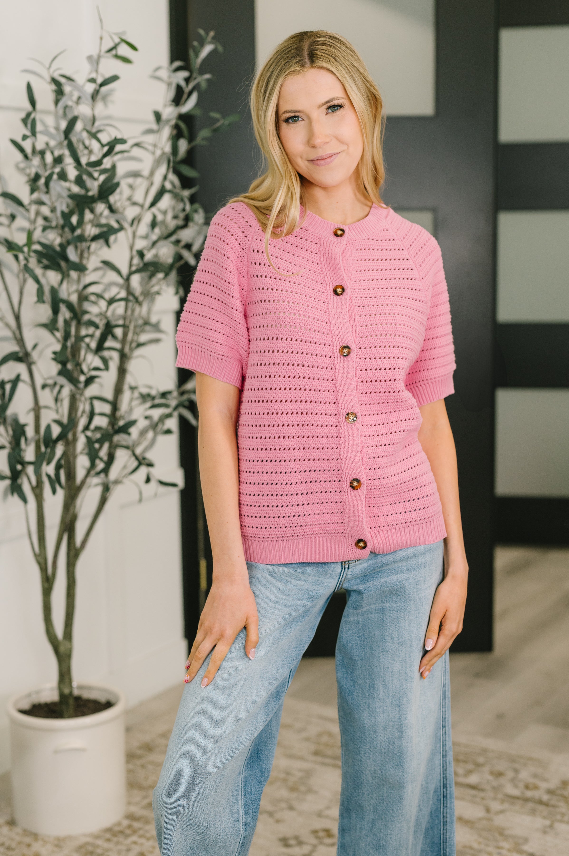 Woman wearing a pink short sleeve knit button down sweater with a round neckline and light wash wide leg jeans indoors